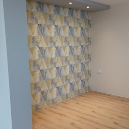 Marcin Polek Usługi Remontowo-Serwisowe MM - Pusty pokój po remoncie z drewnianą podłogą i ścianą pokrytą tapetą w geometryczne wzory trójkątów w odcieniach szarości i złota, oświetlony punktowymi lampami sufitowymi.