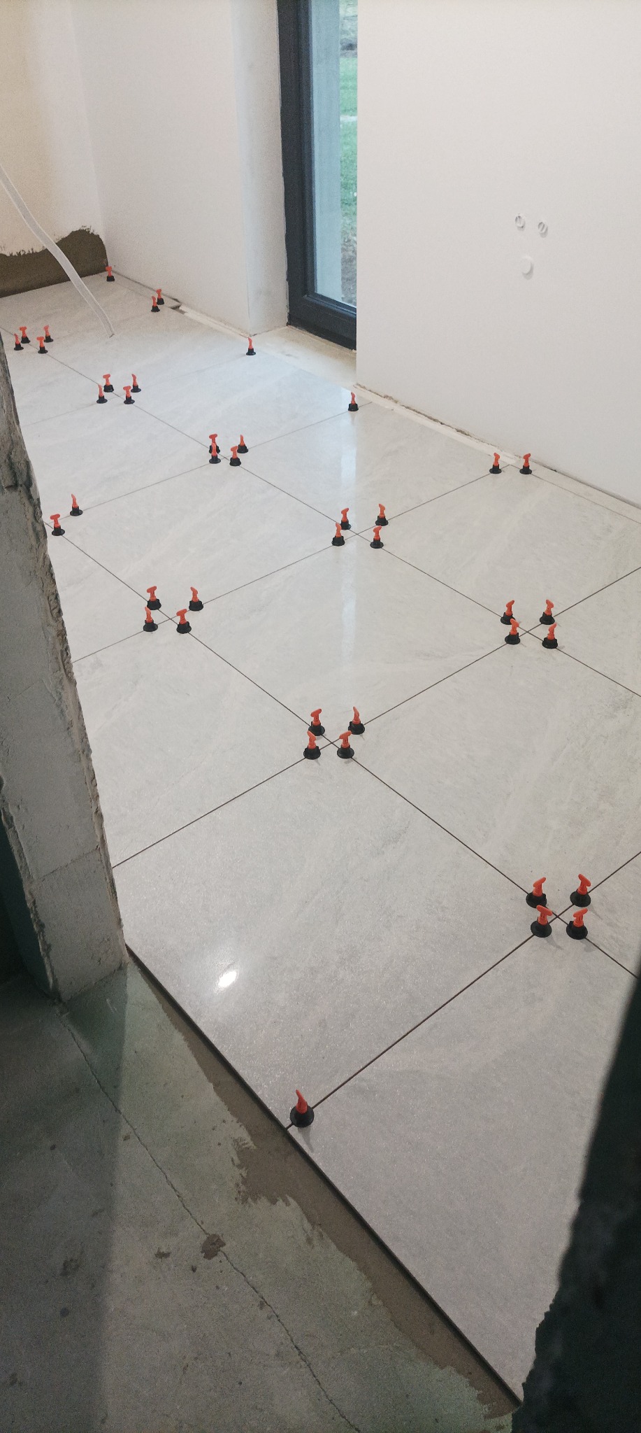 Jasnoszare płytki podłogowe ułożone z użyciem systemu poziomowania płytek (klipsy i kliny), widoczne surowe ściany i okno.