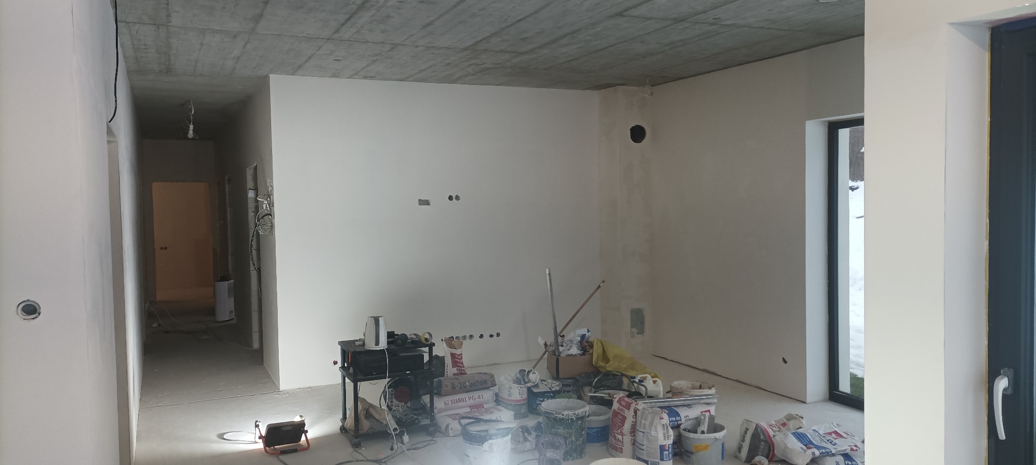 Wnętrze w stanie deweloperskim w Bochni, ściany po nałożeniu gładzi, widoczne worki z zaprawą, narzędzia i prowizoryczne oświetlenie podłogowe.