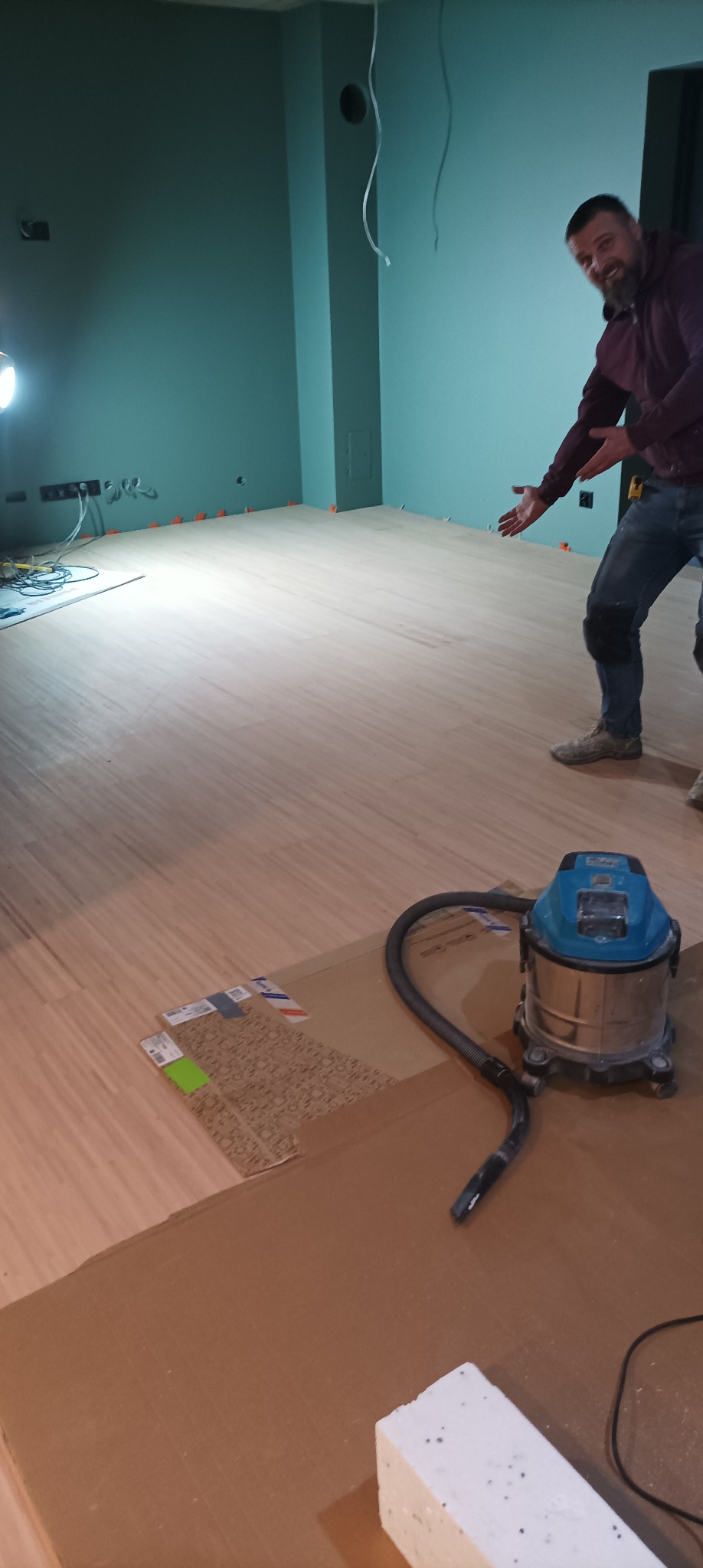 Układanie paneli podłogowych w pomieszczeniu z pomalowanymi na zielono ścianami, widoczny mężczyzna wskazujący na podłogę, odkurzacz przemysłowy i kostka styropianu.