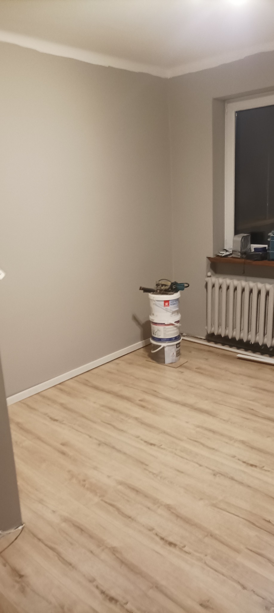 Pomieszczenie po świeżym malowaniu ścian na szaro, z widoczną podłogą imitującą drewno i ustawionymi w rogu trzema wiadrami z farbą oraz narzędziami malarskimi na wierzchu.