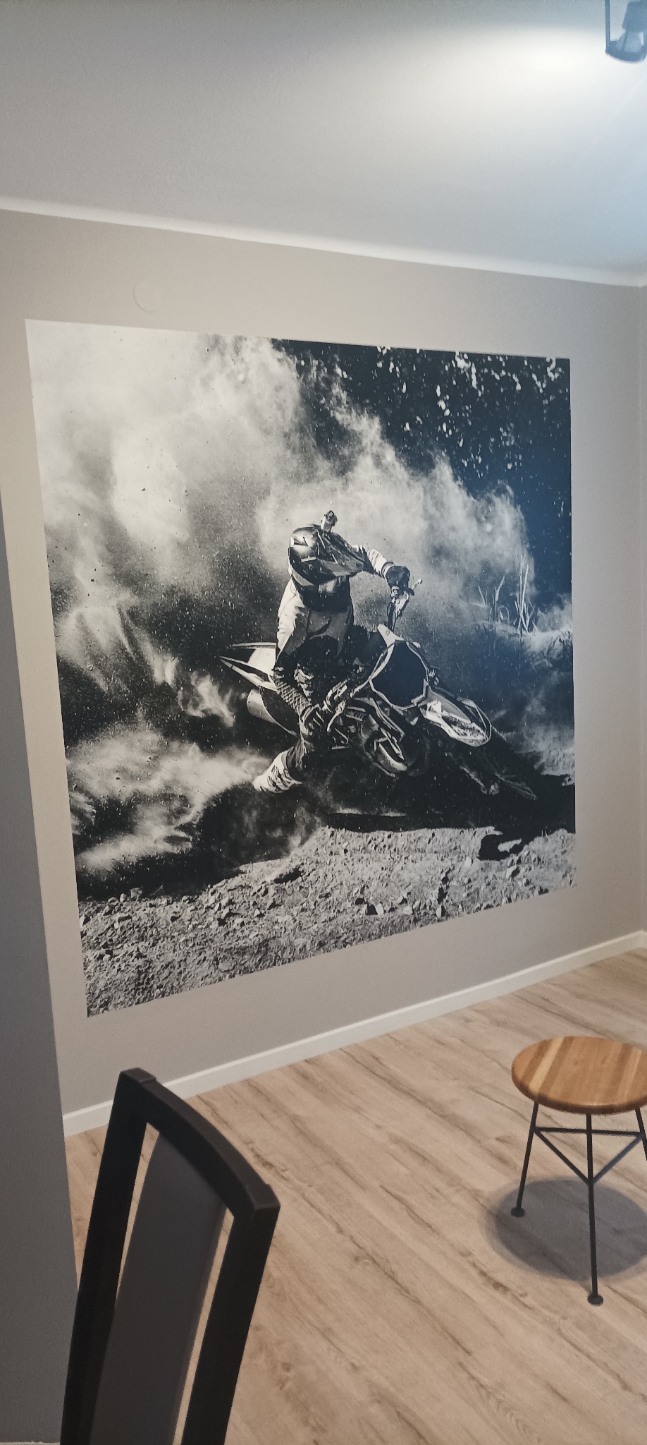 Czarno-biała fototapeta na ścianie przedstawiająca motocyklistę w kasku podczas jazdy w terenie, widoczny fragment krzesła i stołka na tle paneli podłogowych