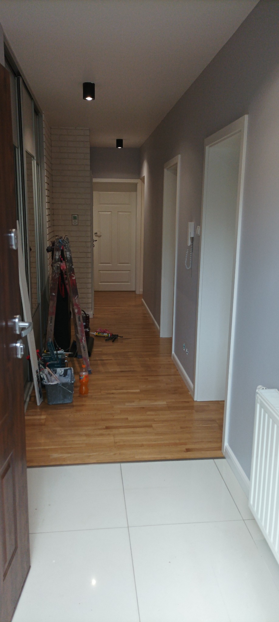 Korytarz w trakcie prac wykończeniowych, widoczna drabina malarska z narzędziami, ściany pomalowane na szaro, drewniana podłoga przechodzi w białe płytki, oświetlenie sufitowe punktowe.