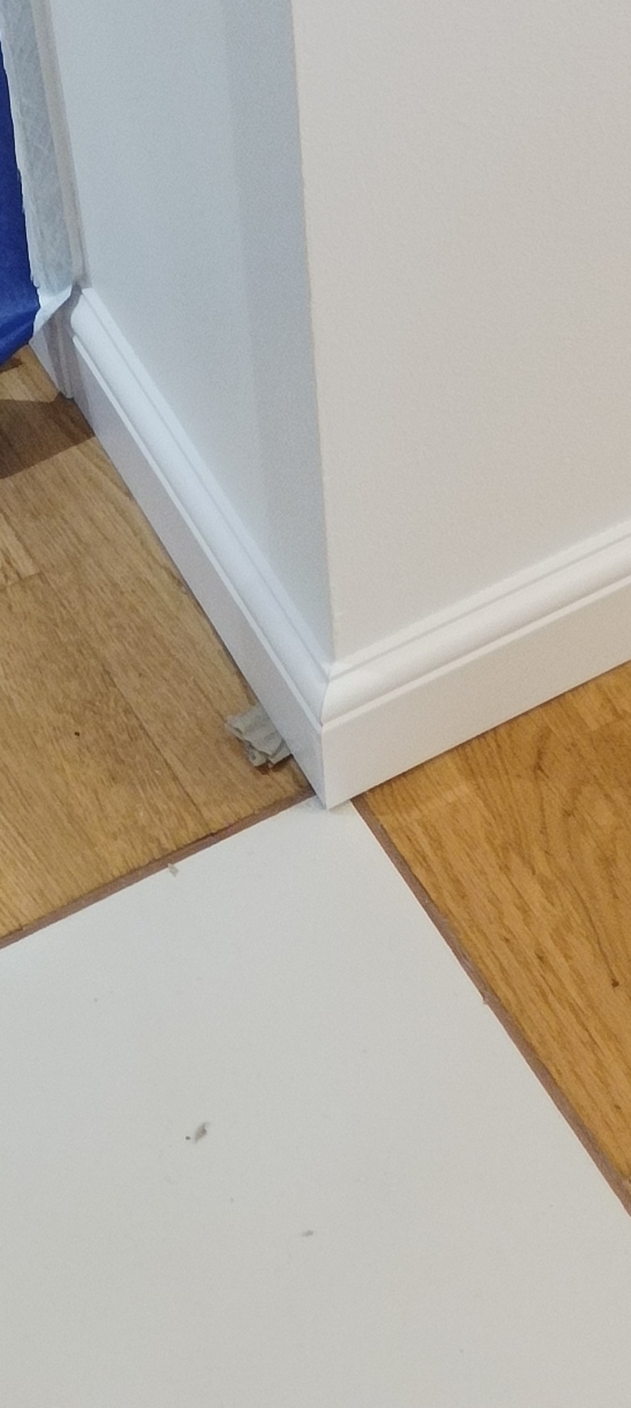 Świeżo pomalowany narożnik ściany z białą listwą przypodłogową, częściowo przykryty folią malarską na drewnianej podłodze.