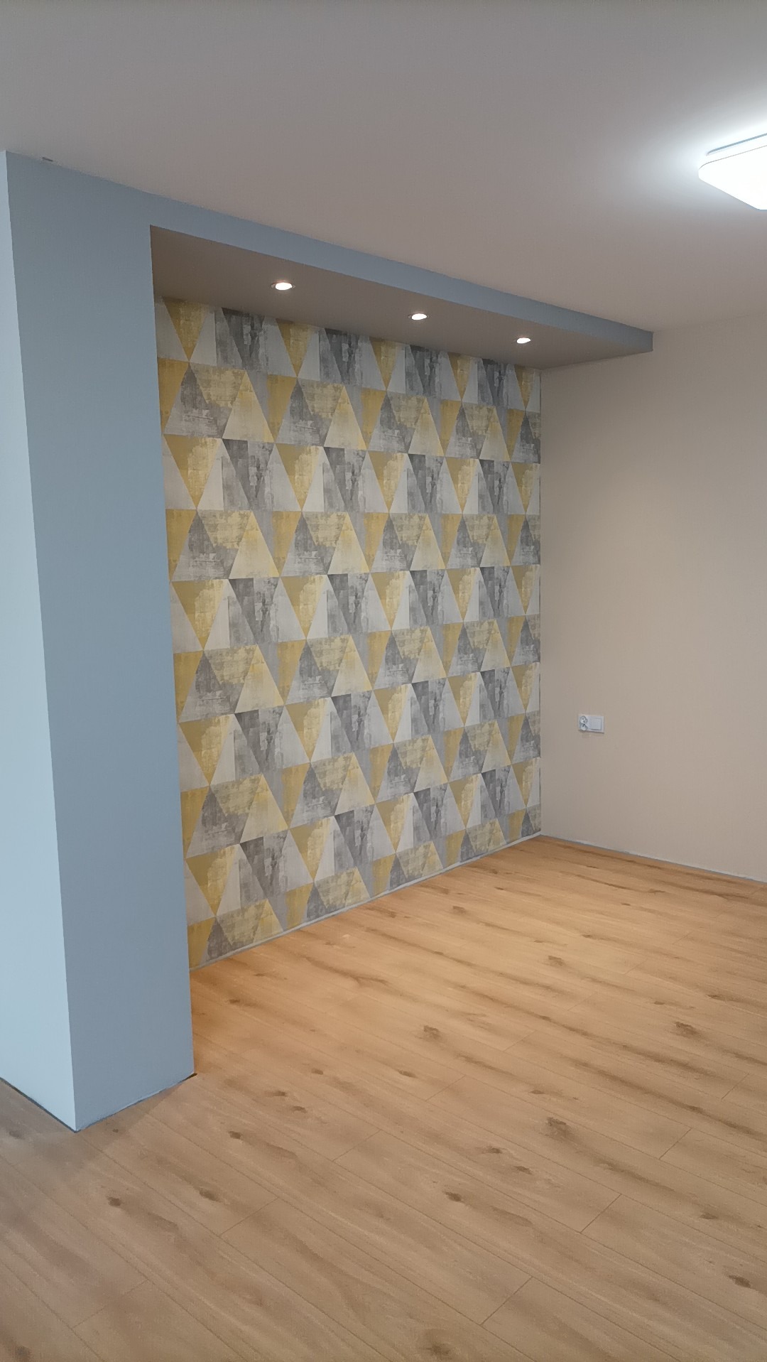 Pusty pokój po remoncie z drewnianą podłogą i ścianą pokrytą tapetą w geometryczne wzory trójkątów w odcieniach szarości i złota, oświetlony punktowymi lampami sufitowymi.