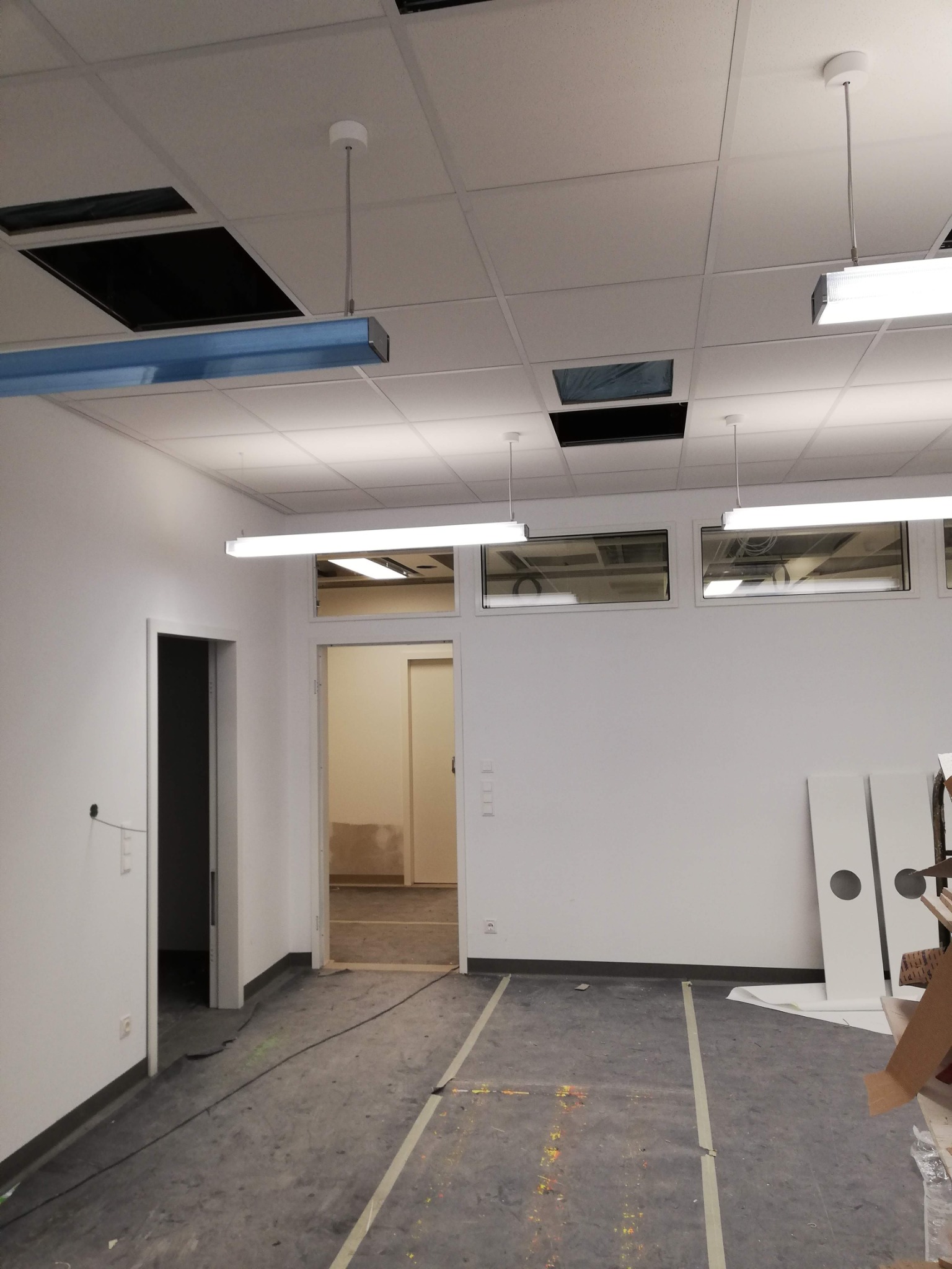 Wnętrze biura w trakcie remontu, widoczne przygotowane ściany pomalowane na biało, sufit podwieszany z otworami, podłoga zabezpieczona taśmą, oraz elementy oświetlenia