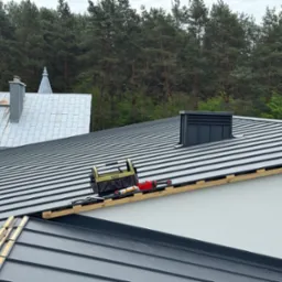 Szary dach z blachy na budynku, z widocznymi narzędziami dekarskimi i fragmentem lasu w tle. Widoczne elementy obróbek blacharskich.