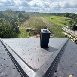 BUD-DACH PAWEŁ GAWROŃSKI - Nowy dach z czarnej dachówki ceramicznej, widok z góry na komin i krajobraz. Na tle zielone pola i błękitne niebo z chmurami.