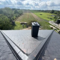 Nowy dach z czarnej dachówki ceramicznej, widok z góry na komin i krajobraz. Na tle zielone pola i błękitne niebo z chmurami.