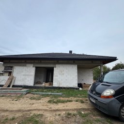 BUD-DACH PAWEŁ GAWROŃSKI - Surowy stan domu jednorodzinnego z czarnym dachem, widok z zewnątrz na tle pochmurnego nieba i zaparkowanego samochodu dostawczego.