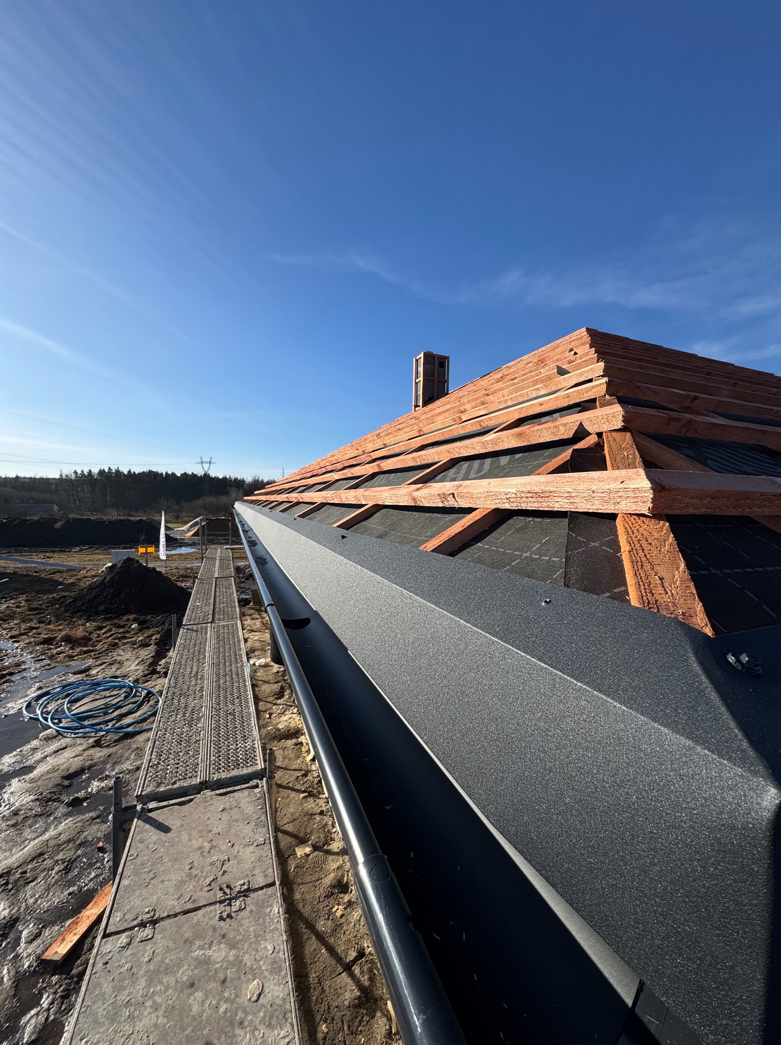 Szara rynna zamontowana na nowym dachu z drewnianą konstrukcją i czarną membraną, widok z rusztowania w słoneczny dzień.