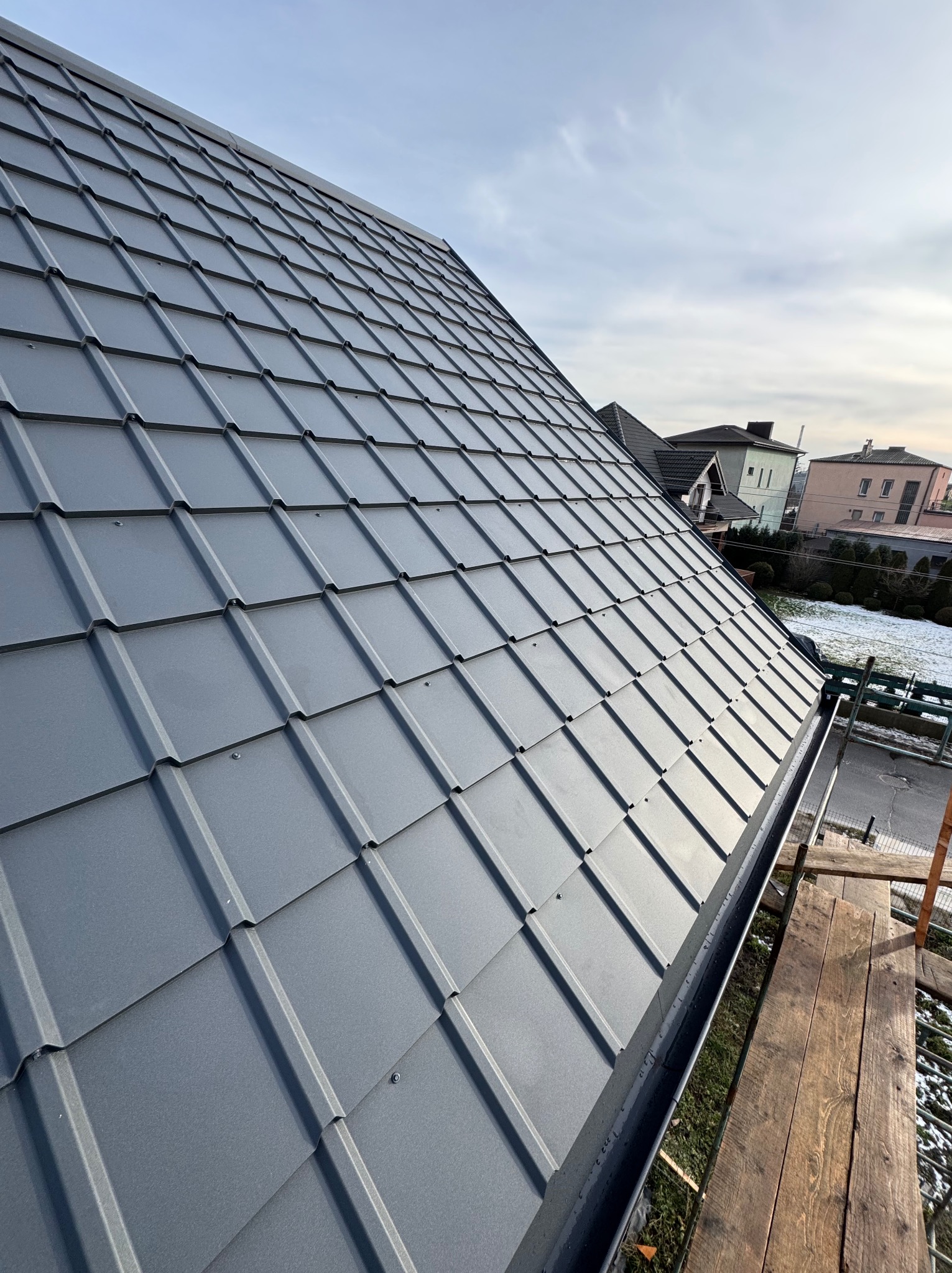 Nowy, grafitowy dach z blachodachówki modułowej, z widoczną rynną, zamontowany na budynku z sąsiedztwem domów jednorodzinnych i resztkami śniegu.