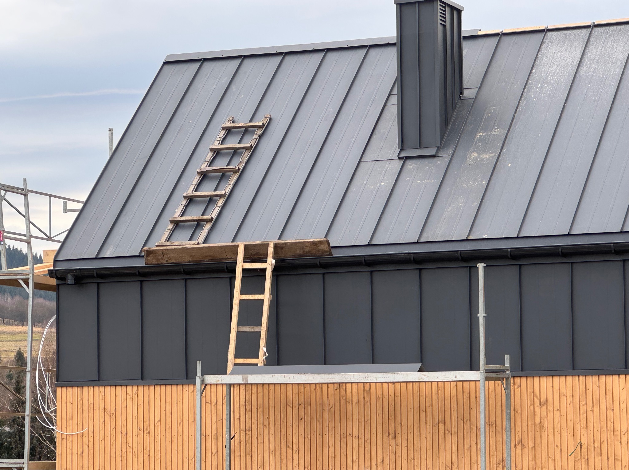 Nowy dach z blachy na rąbek stojący, drewniana drabina oparta o połać, widoczne rusztowanie i komin, elewacja z desek w dwóch odcieniach drewna.