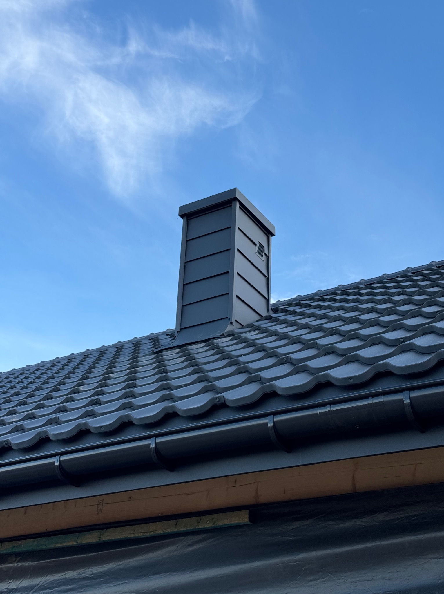 Grafitowy dach z ceramiczną dachówką karpiówką i nowoczesnym kominem na tle błękitnego nieba z chmurami.