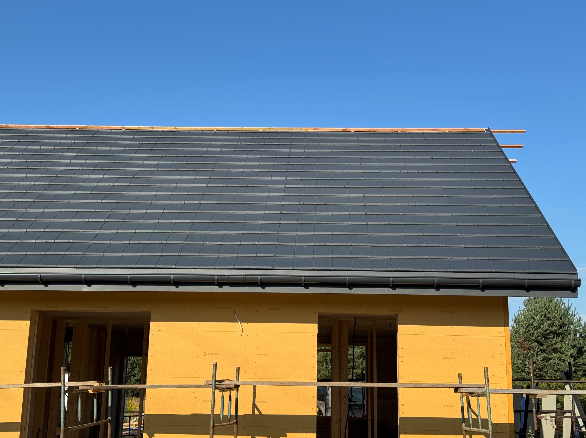 Nowy, ciemny dach z rynnami na budynku w trakcie budowy, widok z dołu na tle błękitnego nieba. Widoczne ocieplenie ścian budynku.