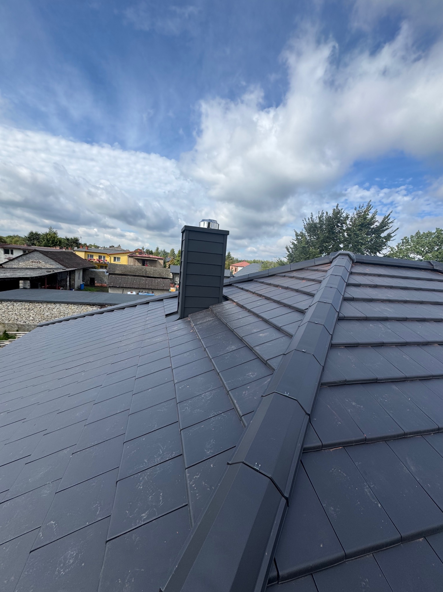 Nowy, ciemnoszary dach z kominem na tle błękitnego nieba z chmurami. Widok z góry, ukazujący regularny wzór dachówek i obróbki blacharskie.