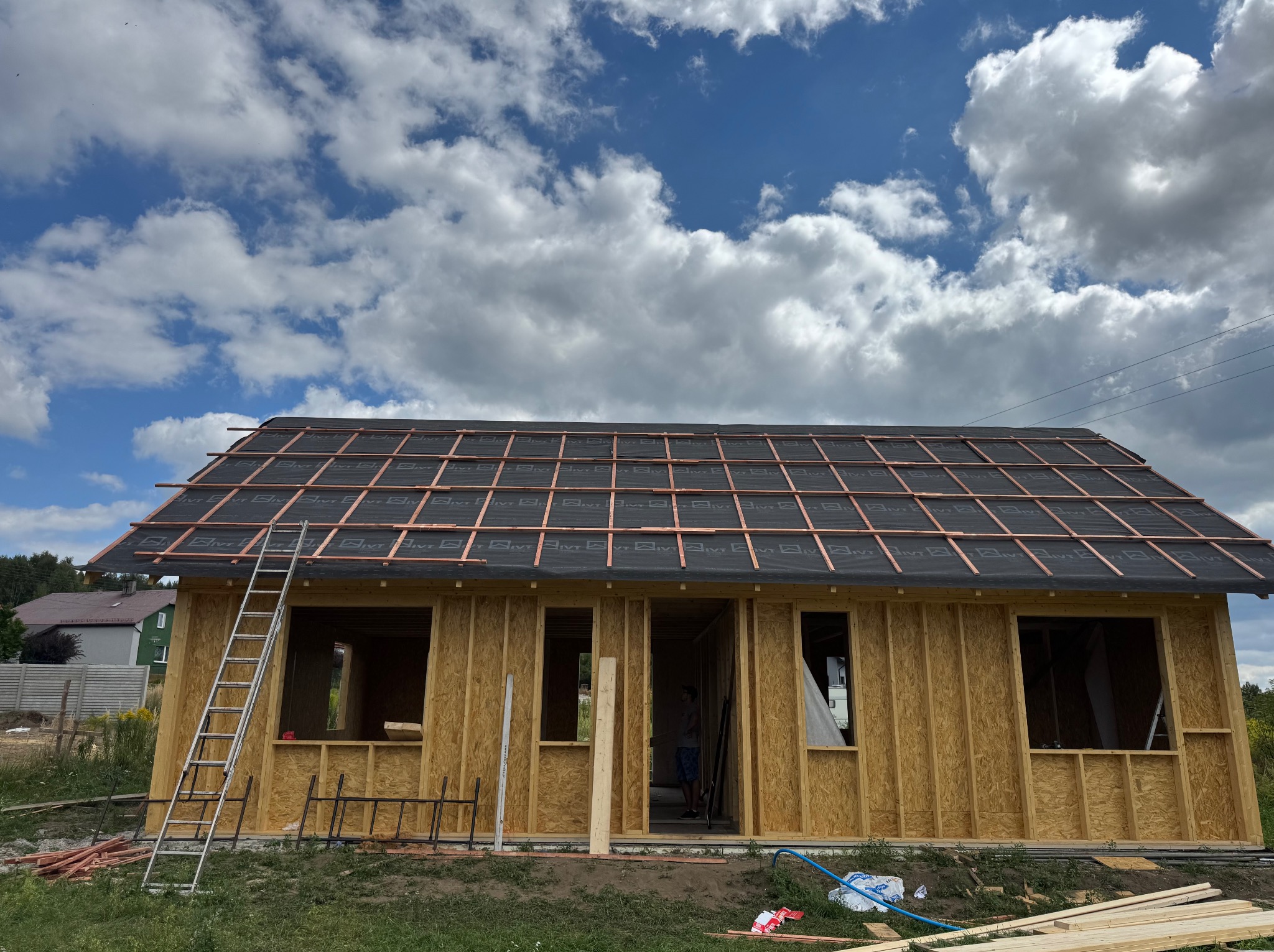 Budowa domu w trakcie: drewniana konstrukcja z deskami, pokrycie dachu membraną i łatami, drabina oparta o ścianę. Błękitne niebo z obłokami w tle.