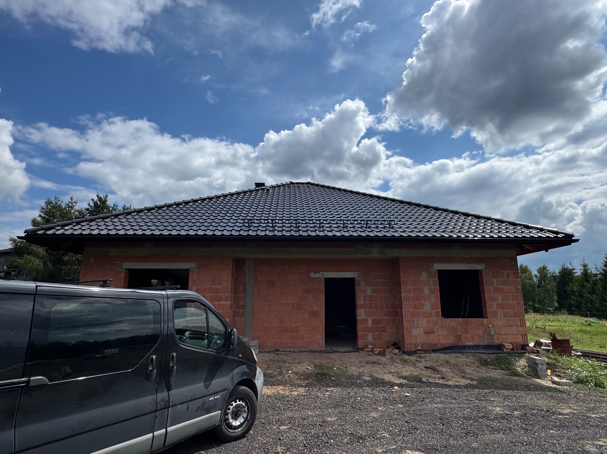 Dom w trakcie budowy z czarnym, błyszczącym dachem ceramicznym na tle błękitnego nieba z chmurami. Częściowo widoczny samochód dostawczy z lewej strony.