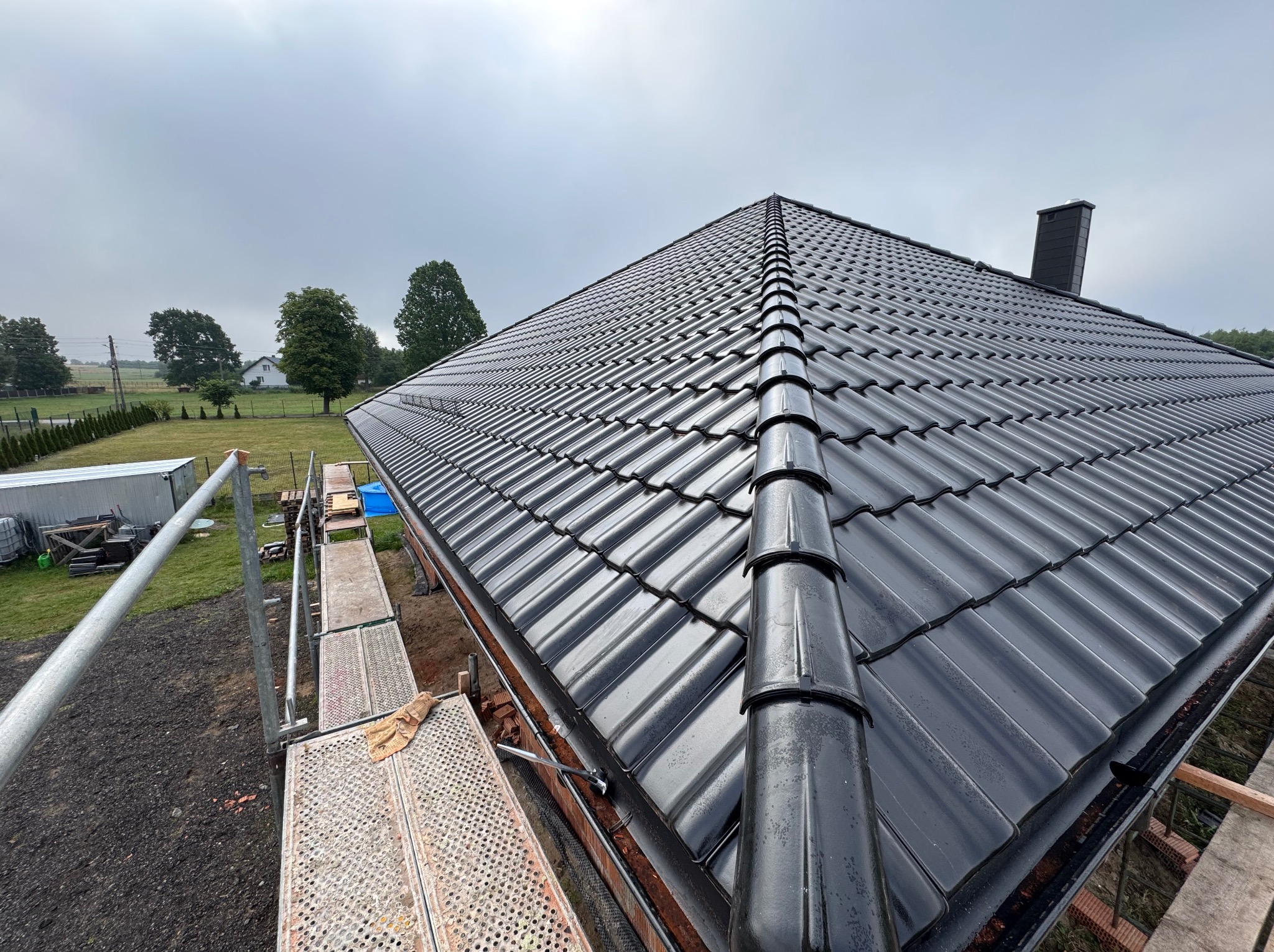 Błyszczący, czarny dach z dachówki ceramicznej widziany z rusztowania, z kominem w tle i zielonym otoczeniem. Widok z góry na wykończony dach.