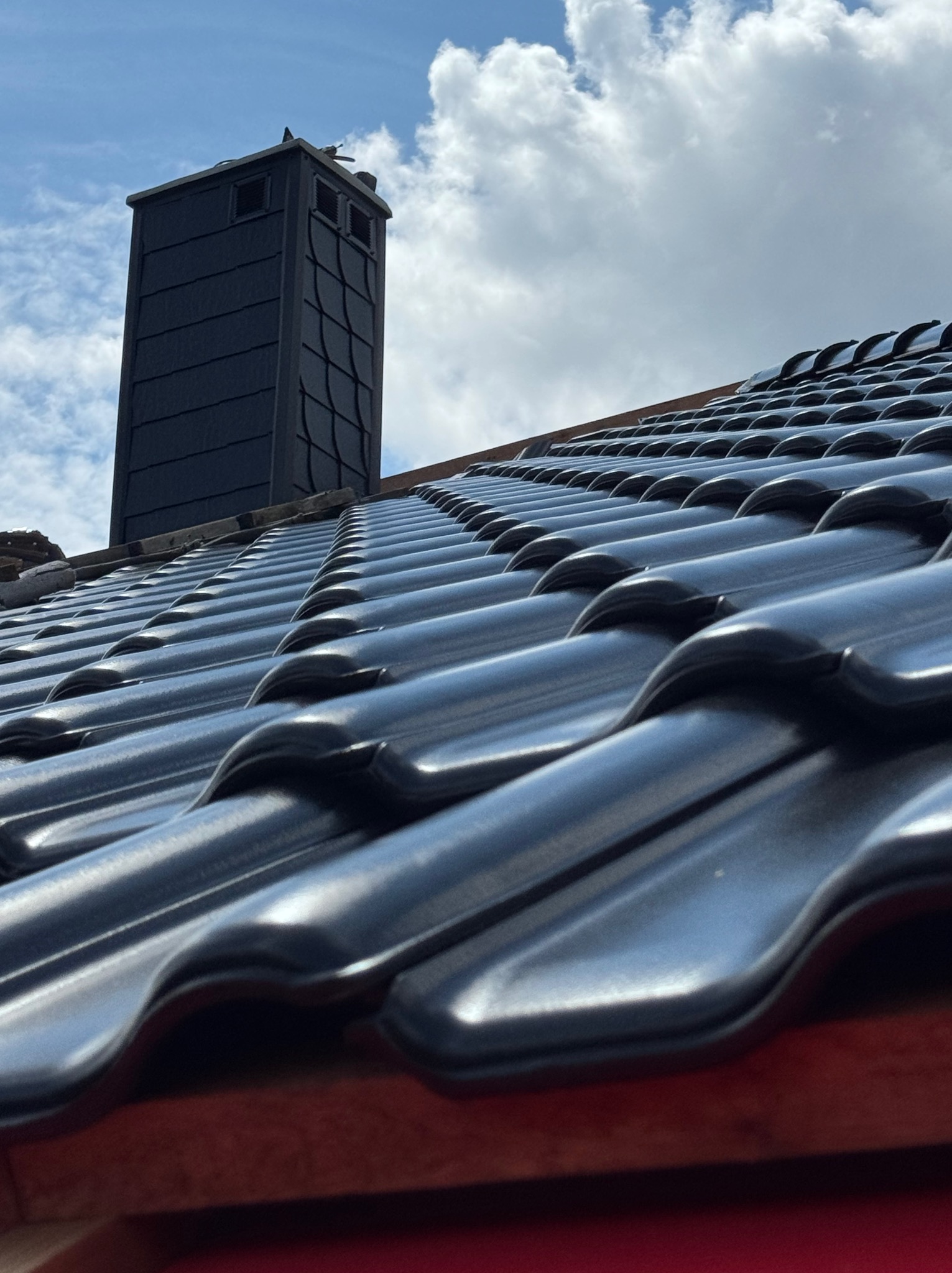 Błyszcząca, czarna dachówka ceramiczna na skośnym dachu, z kominem w tle, na tle pochmurnego nieba. Widok z dołu, ukazujący regularny wzór dachówek.