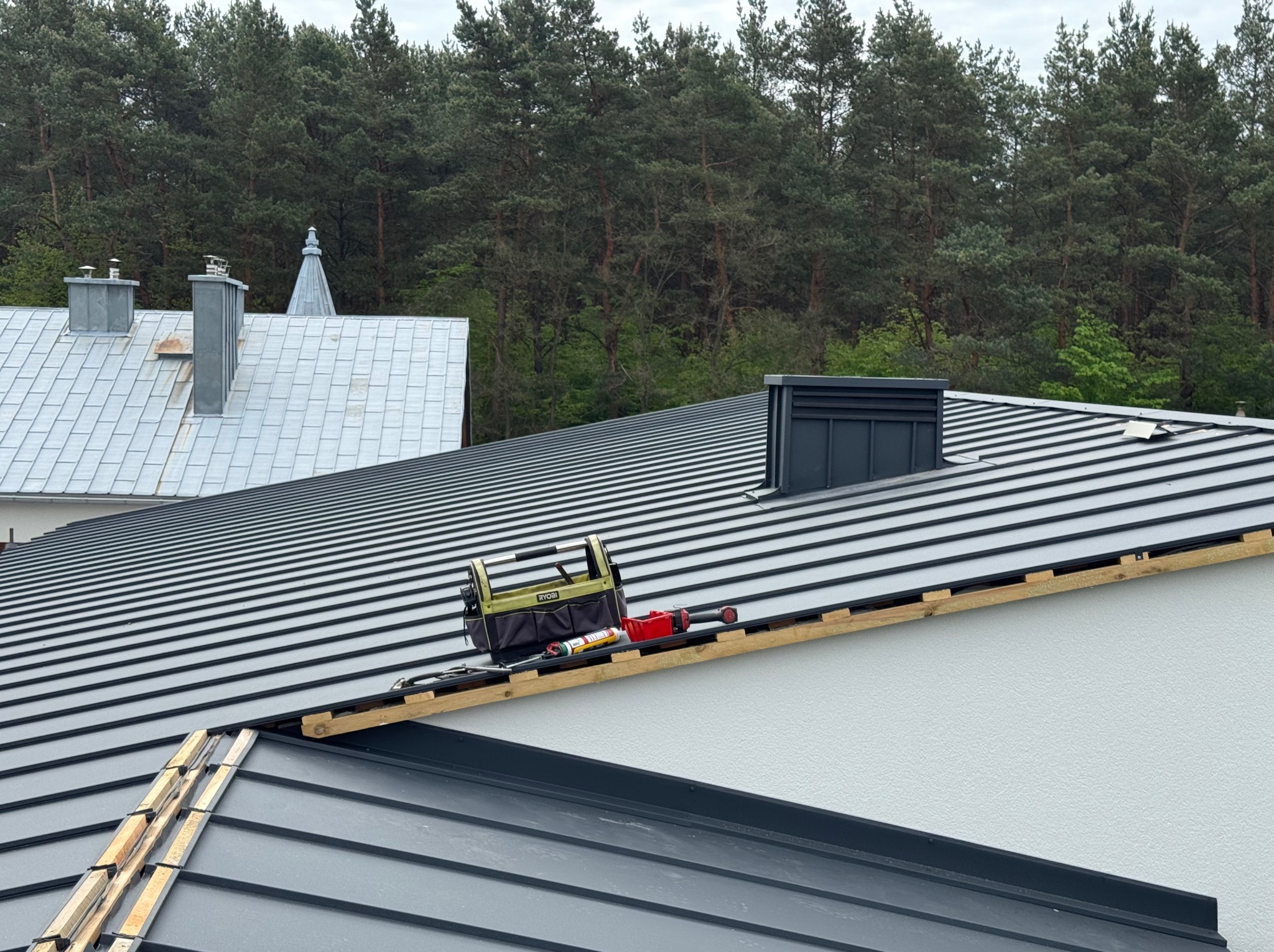 Szary dach z blachy na budynku, z widocznymi narzędziami dekarskimi i fragmentem lasu w tle. Widoczne elementy obróbek blacharskich.