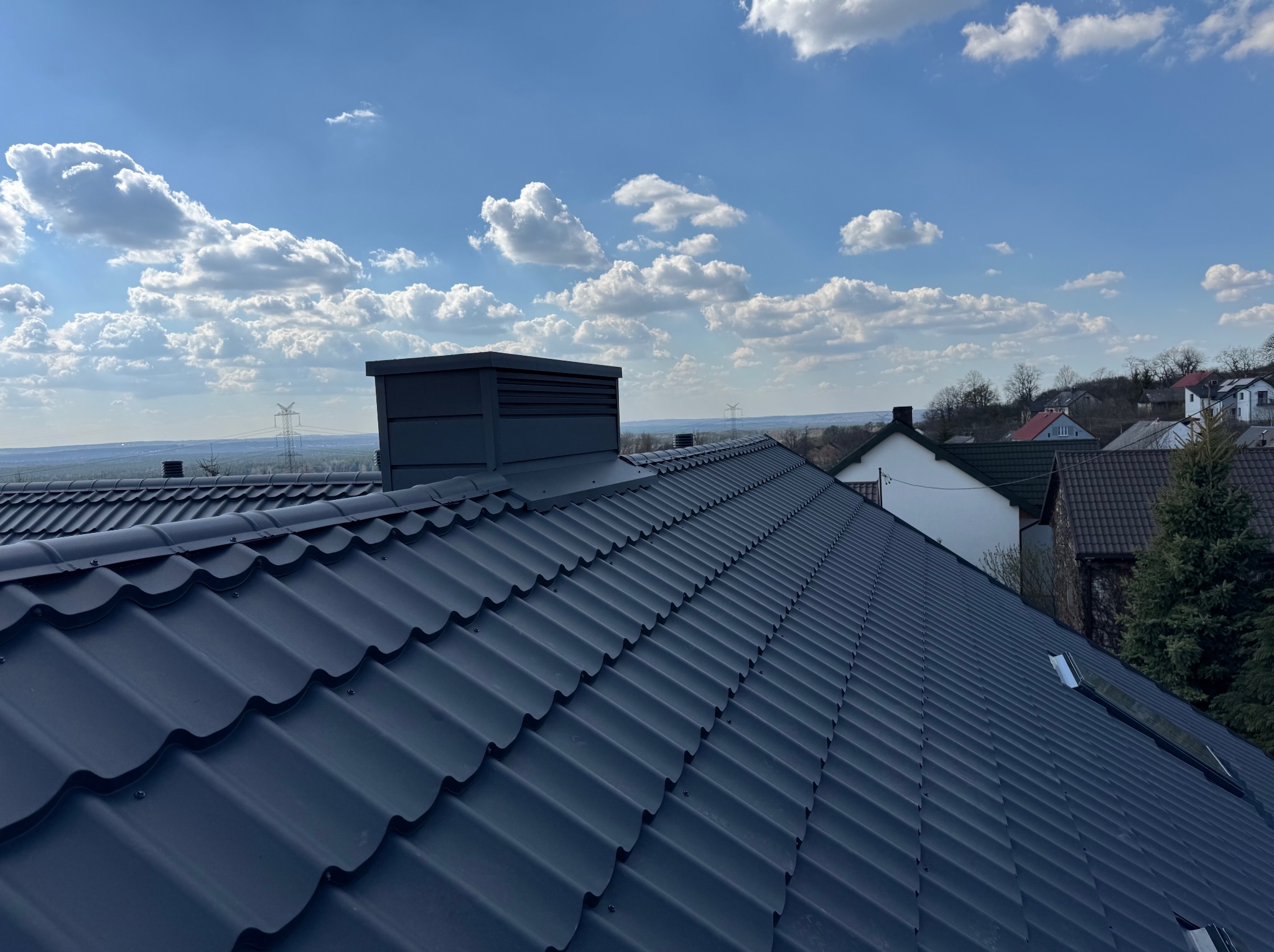 Nowy, ciemny dach z blachy trapezowej, widok z góry, komin wentylacyjny, błękitne niebo z chmurami w tle. Widoczne domy w oddali. Dach po remoncie.