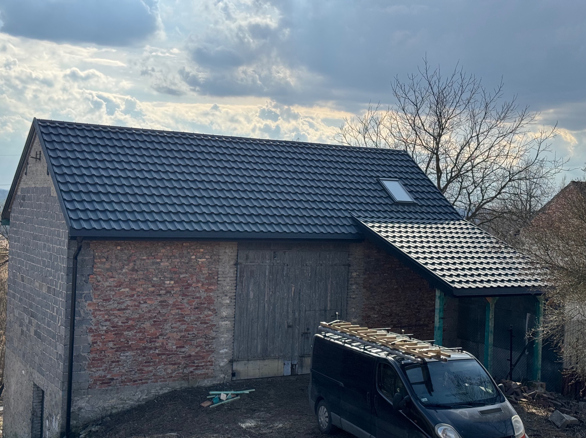 Dom z nowym, grafitowym dachem z blachodachówki i oknem dachowym. Na dachu zaparkowany czarny van z drabiną. Część ściany budynku z czerwonej cegły.