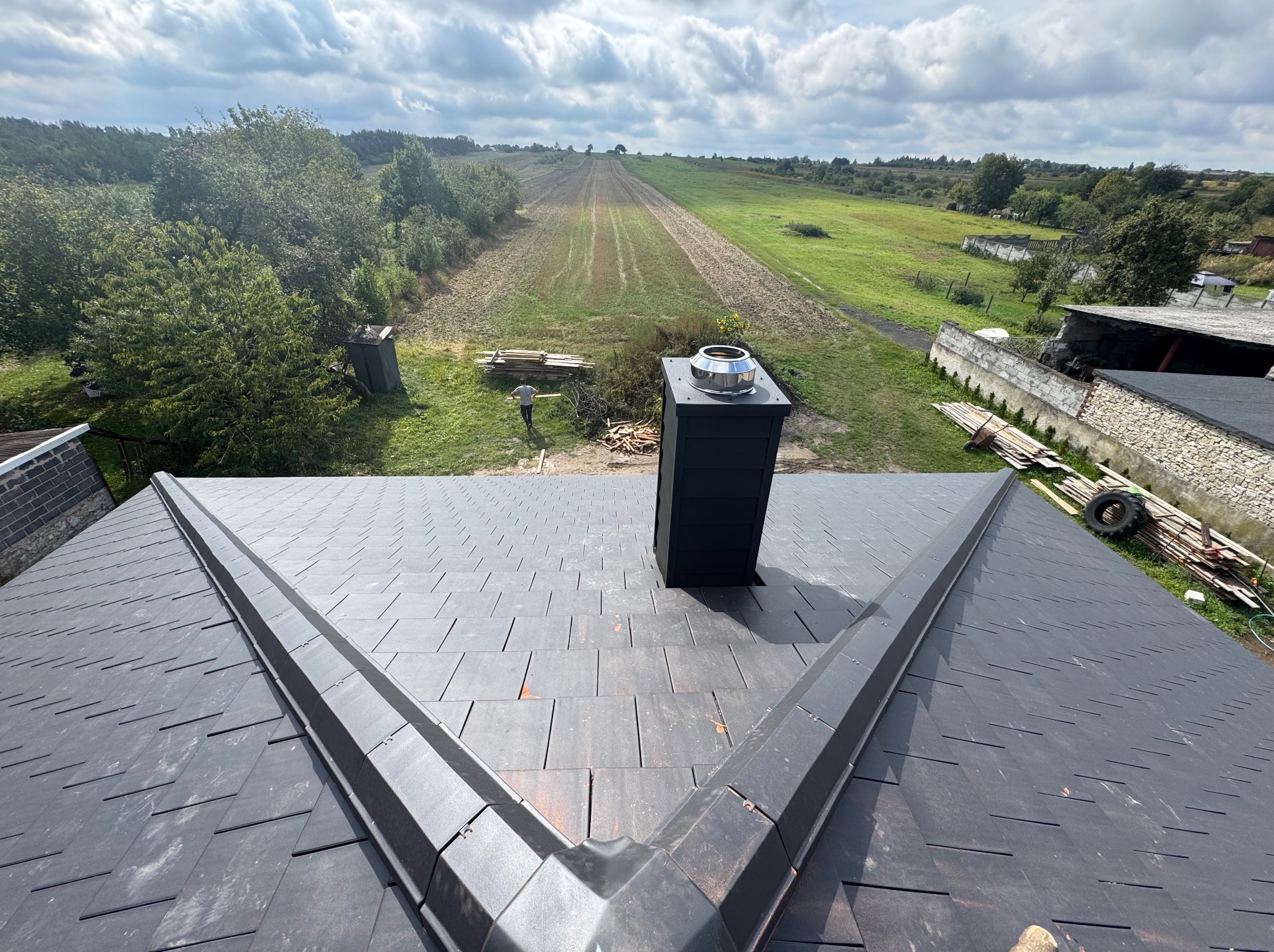 Nowy dach z czarnej dachówki ceramicznej, widok z góry na komin i krajobraz. Na tle zielone pola i błękitne niebo z chmurami.