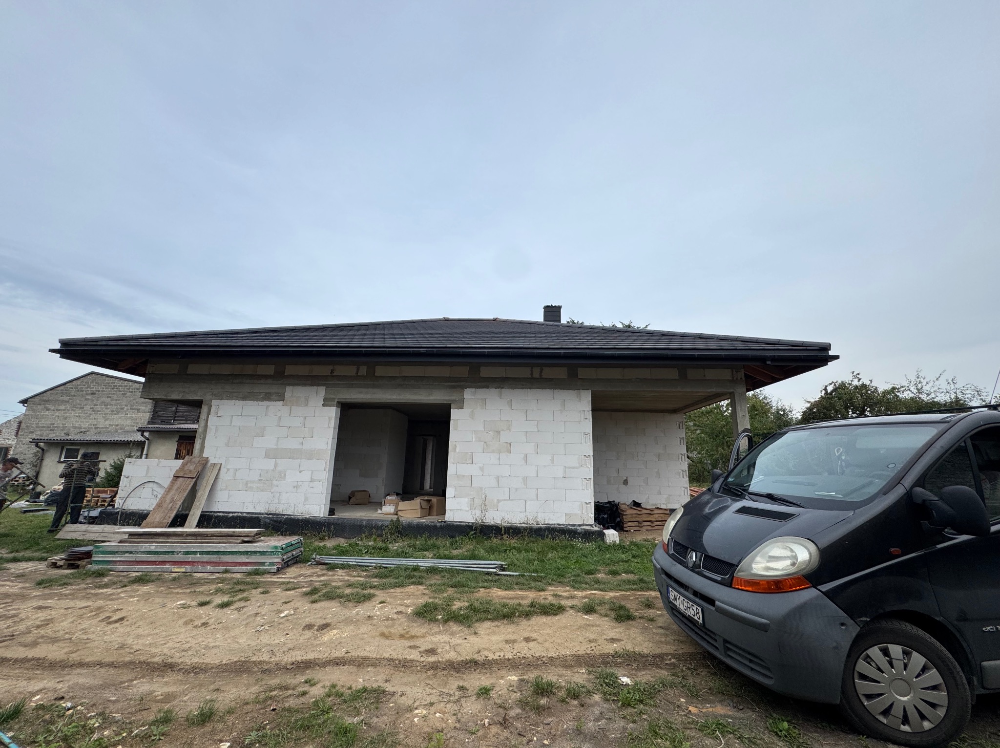 Surowy stan domu jednorodzinnego z czarnym dachem, widok z zewnątrz na tle pochmurnego nieba i zaparkowanego samochodu dostawczego.