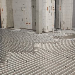 TriTerm Artur Czernik - Białe rury ogrzewania podłogowego ułożone na folii i siatce w surowym wnętrzu z oknami i drabiną. Widok z góry na instalację przed wylaniem posadzki.