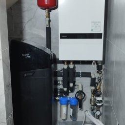 TriTerm Artur Czernik - Zmontowany, nowoczesny piec gazowy Panasonic z czarnym zmiękczaczem wody Clever, czerwonym zbiornikiem wyrównawczym i filtrami wody w pomieszczeniu wyłożonym szarą płytką.