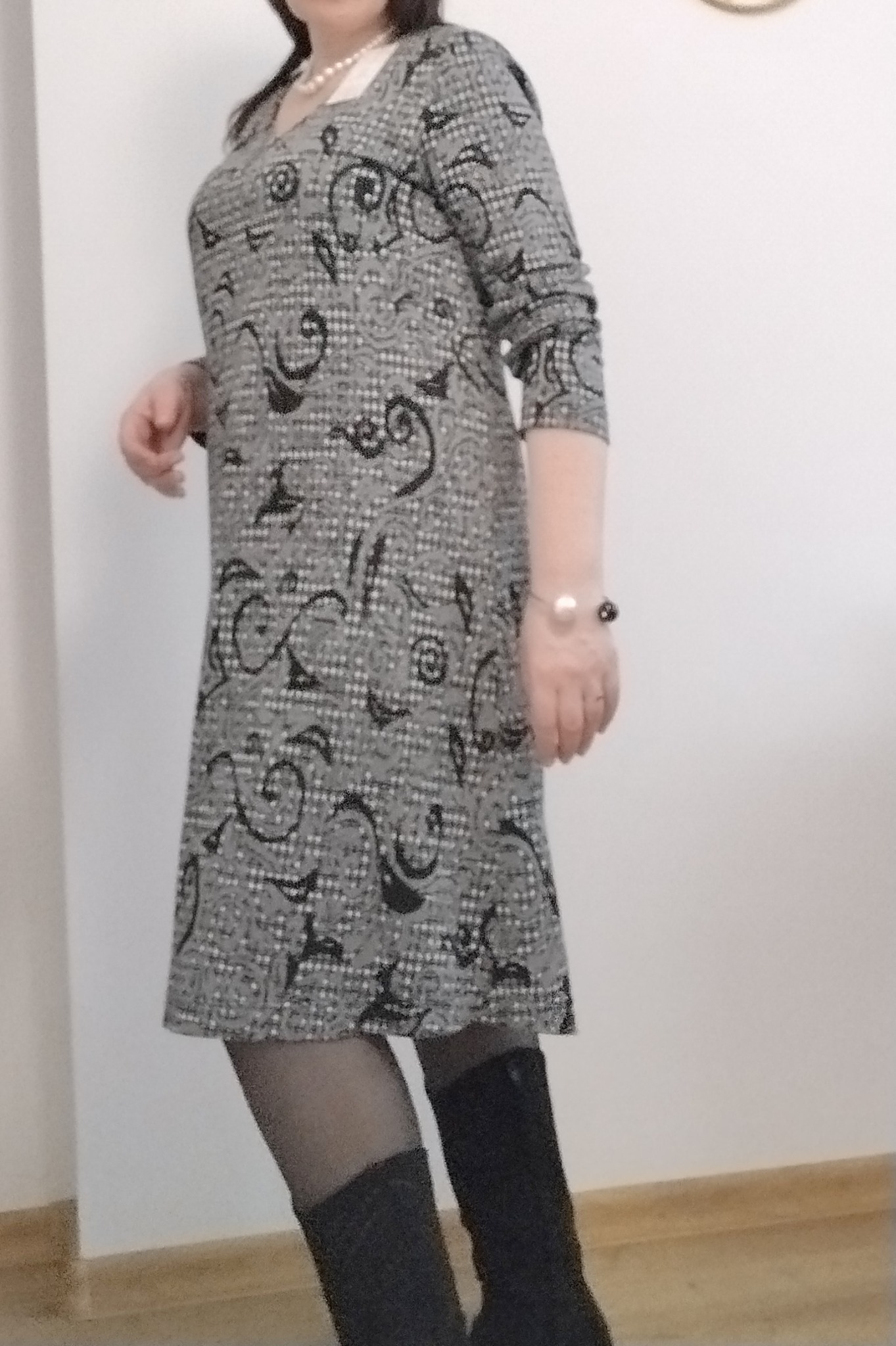 Kobieta pozuje w szarej sukience w czarne wzory, z perłowym naszyjnikiem i bransoletką, czarnych rajstopach i kozakach.