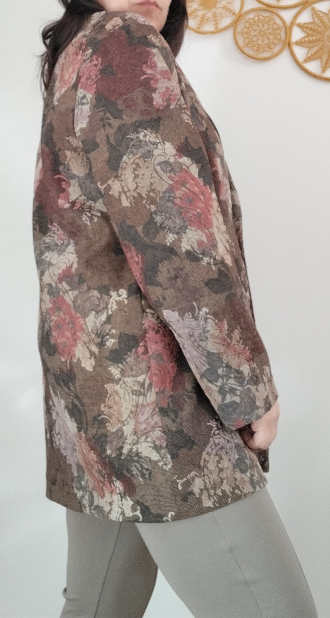 Kobieta w brązowym, wzorzystym żakiecie z motywem kwiatowym, stojąca na tle białej ściany z dekoracjami.