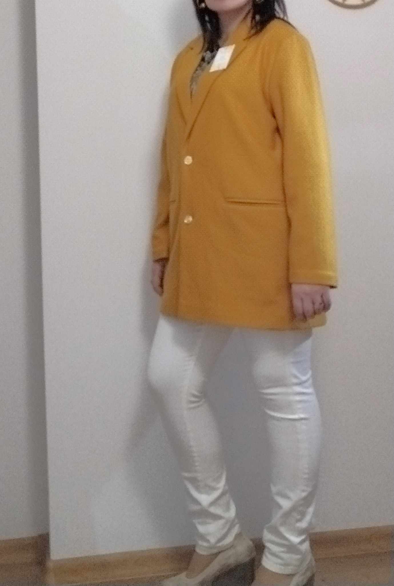 Osoba pozuje w musztardowym, wełnianym żakiecie z klapami, białych spodniach i butach na koturnie. Widoczna metka na żakiecie.