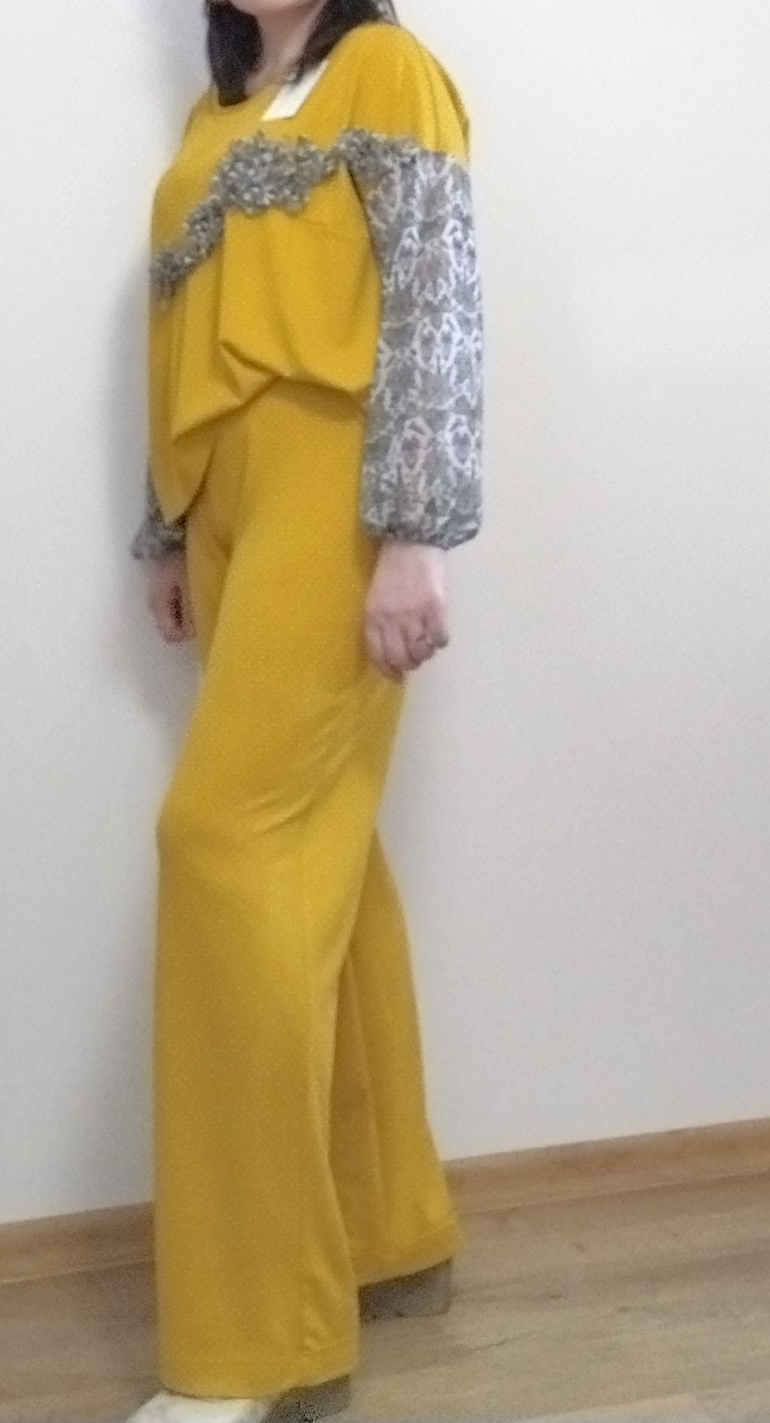 Modelka prezentuje dwuczęściowy, musztardowy komplet odzieży z dekoracyjnymi, koronkowymi rękawami i wstawką na bluzce, na tle jasnej ściany.