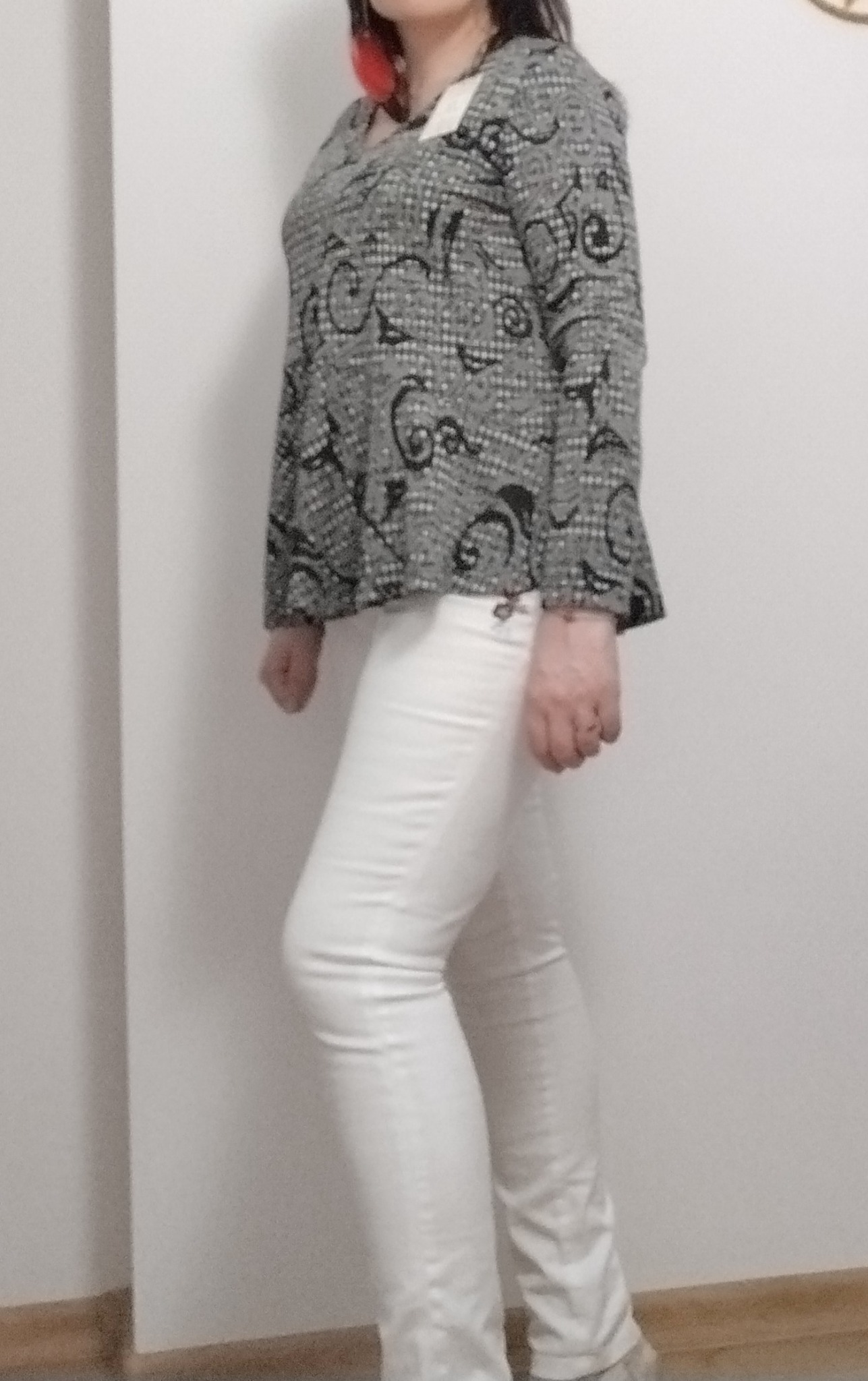 Kobieta prezentuje bluzkę z szaro-czarnym wzorem i białe spodnie, stojąc na tle białej ściany i drewnianej podłogi.