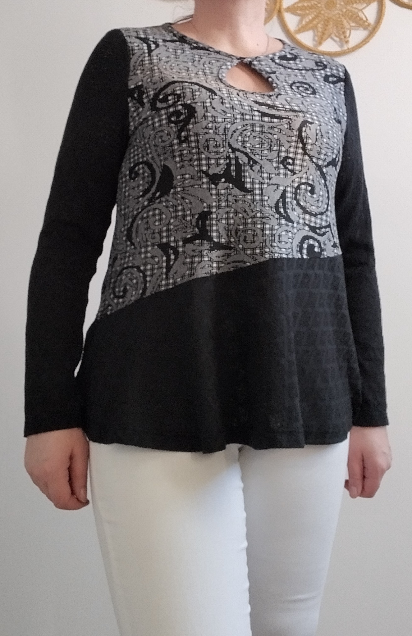 Damska bluzka z długim rękawem, łącząca wzorzystą, szaro-czarną górę z wycięciem na dekolcie i czarnym dołem, zestawiona z białymi spodniami na tle jasnej ściany.