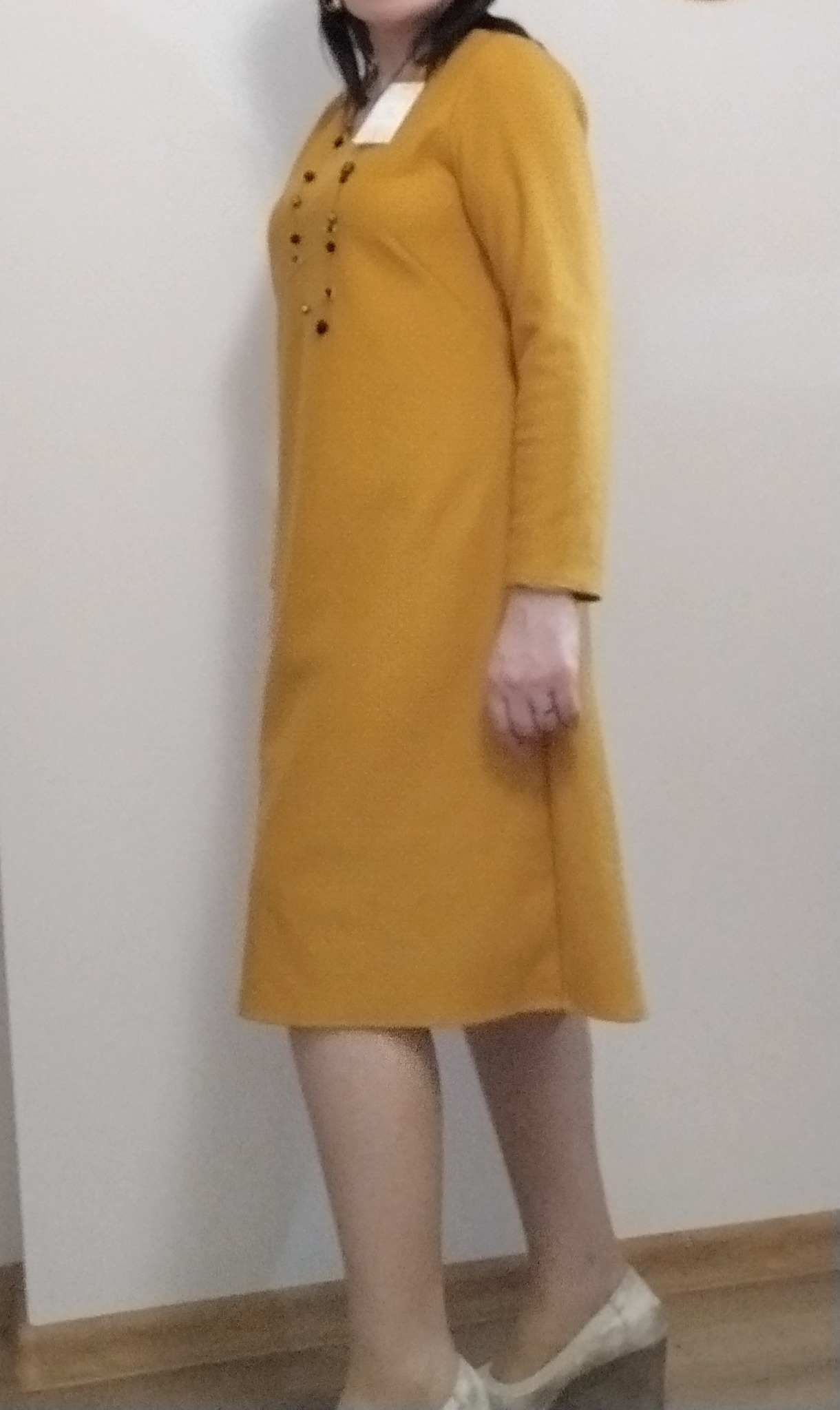 Musztardowa sukienka z długim rękawem i ozdobnymi guzikami, widoczna z boku, z metką przy kołnierzu i butami na koturnie.