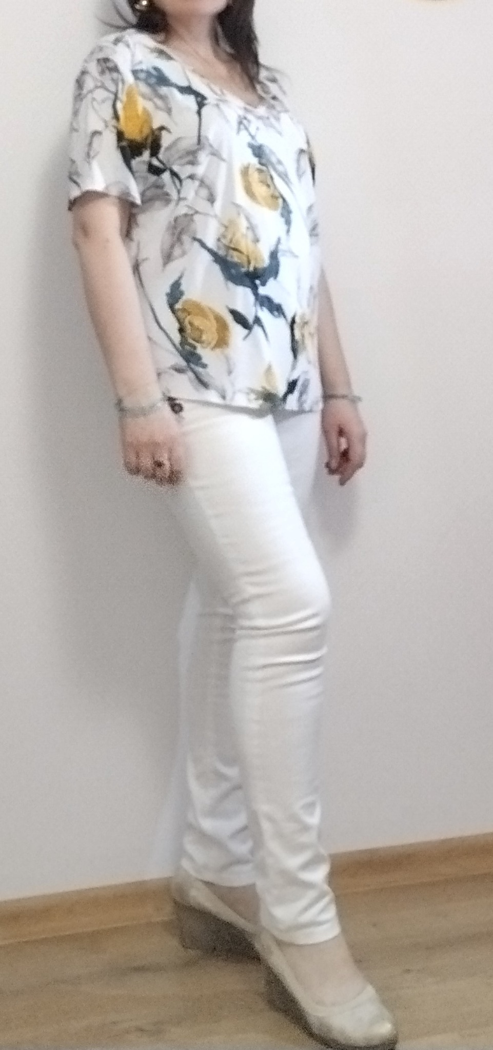 Kobieta pozuje w białych, dopasowanych spodniach i bluzce w kwiaty z krótkim rękawem, uzupełnona srebrnymi bransoletkami i złotymi kolczykami.