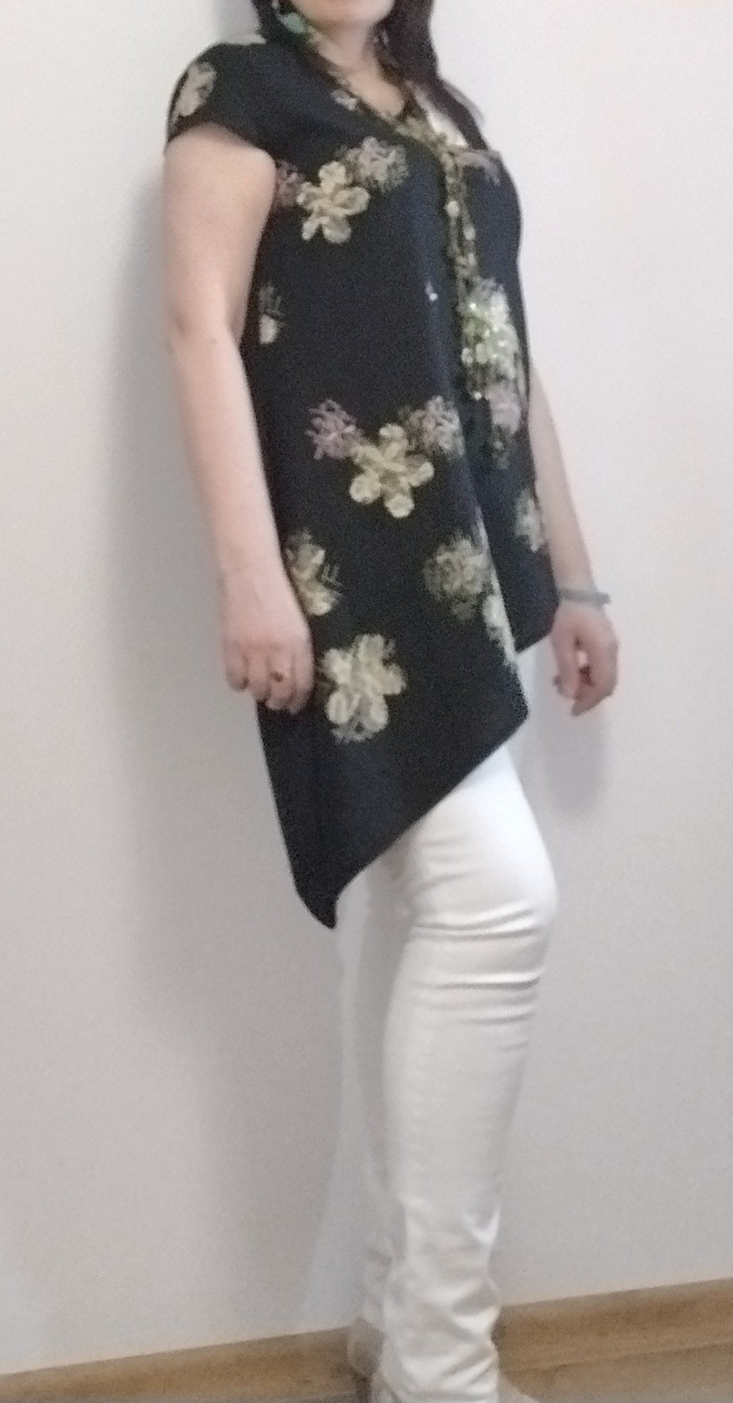 Kobieta pozuje w asymetrycznej tunice w kwiaty i białych spodniach, uzupełnionej o długi naszyjnik z koralików i kolczyki.