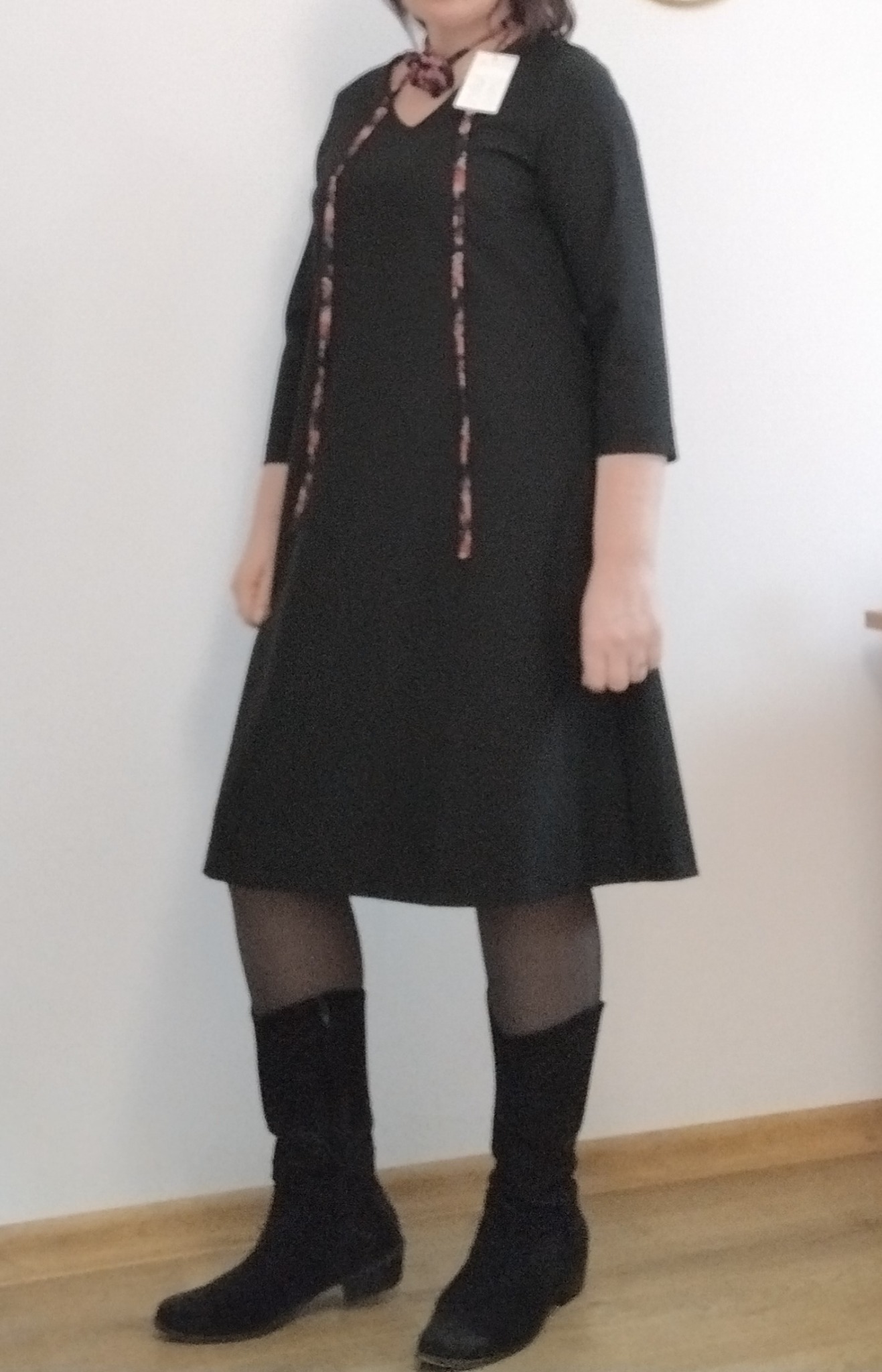 Czarna sukienka z rękawem 3/4 i ozdobnymi sznurkami, prezentowana na modelce w czarnych rajstopach i zamszowych botkach na tle białej ściany.
