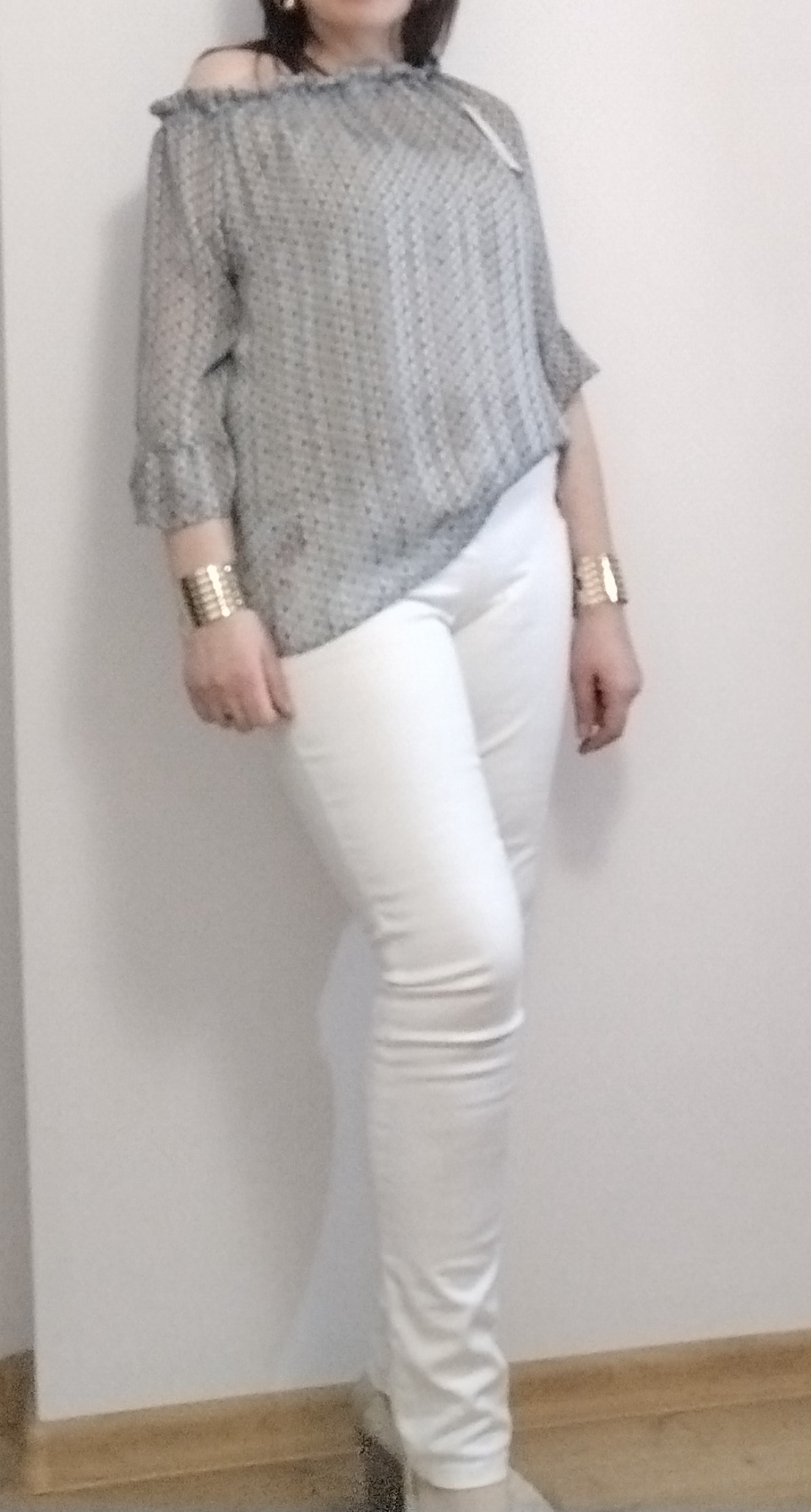 Kobieta pozuje w bluzce z odkrytymi ramionami w szaro-białe wzory i białych, dopasowanych spodniach, z bransoletkami na obu nadgarstkach.