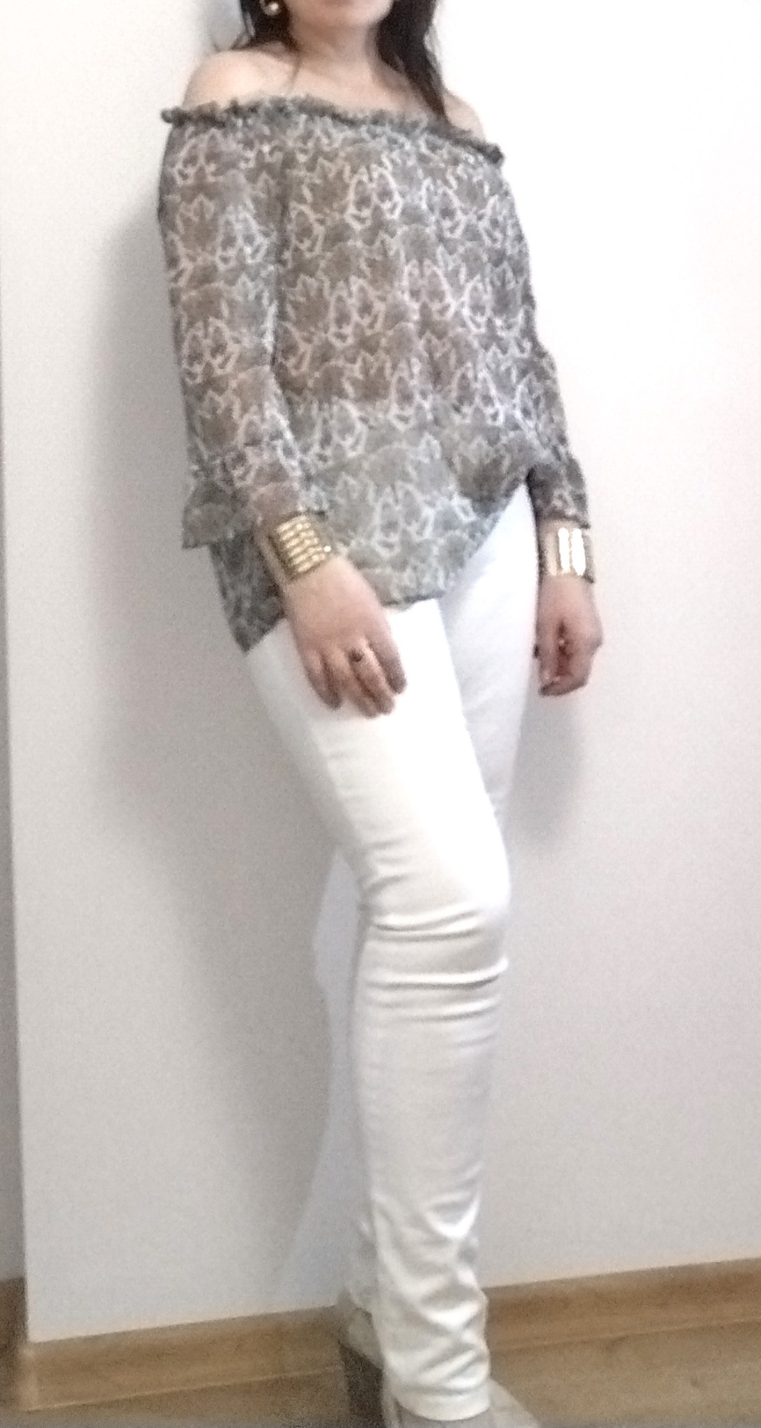 Kobieta pozuje w bluzce z odkrytymi ramionami w szaro-białe wzory i białych, obcisłych spodniach, z grubymi złotymi bransoletkami na obu nadgarstkach.