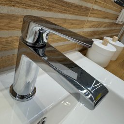 Profesjonalna Firma Sprzątająca - Martyna Zasada - Błyszcząca, chromowana bateria umywalkowa na białej umywalce, z drewnopodobną okładziną ścienną w tle. Widoczne akcesoria łazienkowe.