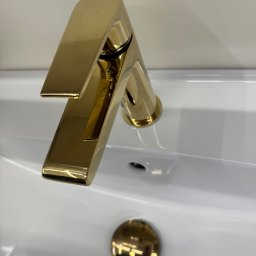Profesjonalna Firma Sprzątająca - Martyna Zasada - Błyszcząca, złota bateria umywalkowa w minimalistycznej, białej umywalce. Nowoczesny design z wyraźnym odbiciem światła.