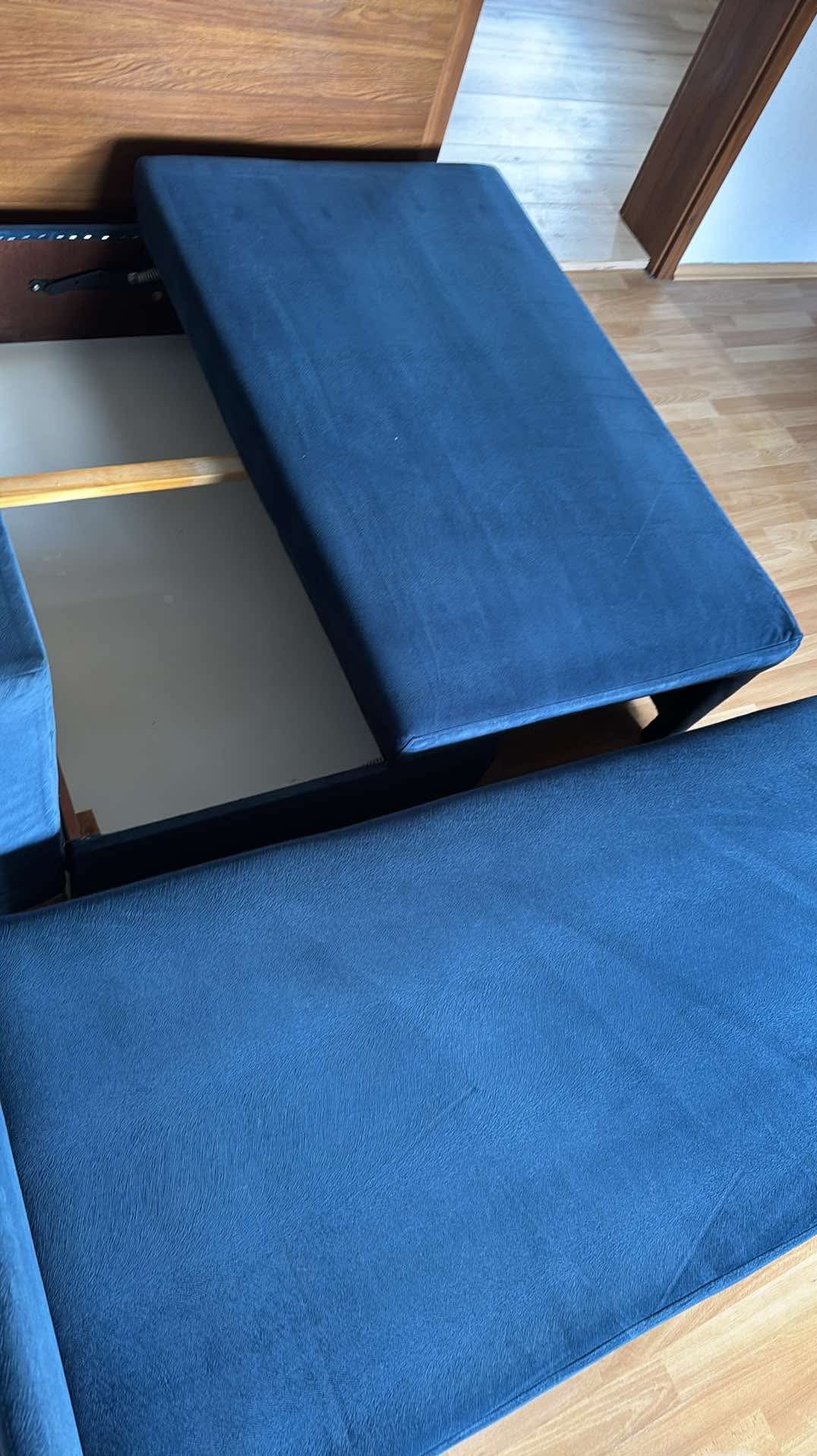 Rozkładana sofa z otwartym pojemnikiem na pościel, tapicerowana granatowym materiałem. Widoczny drewniany korpus i podłoga z paneli.