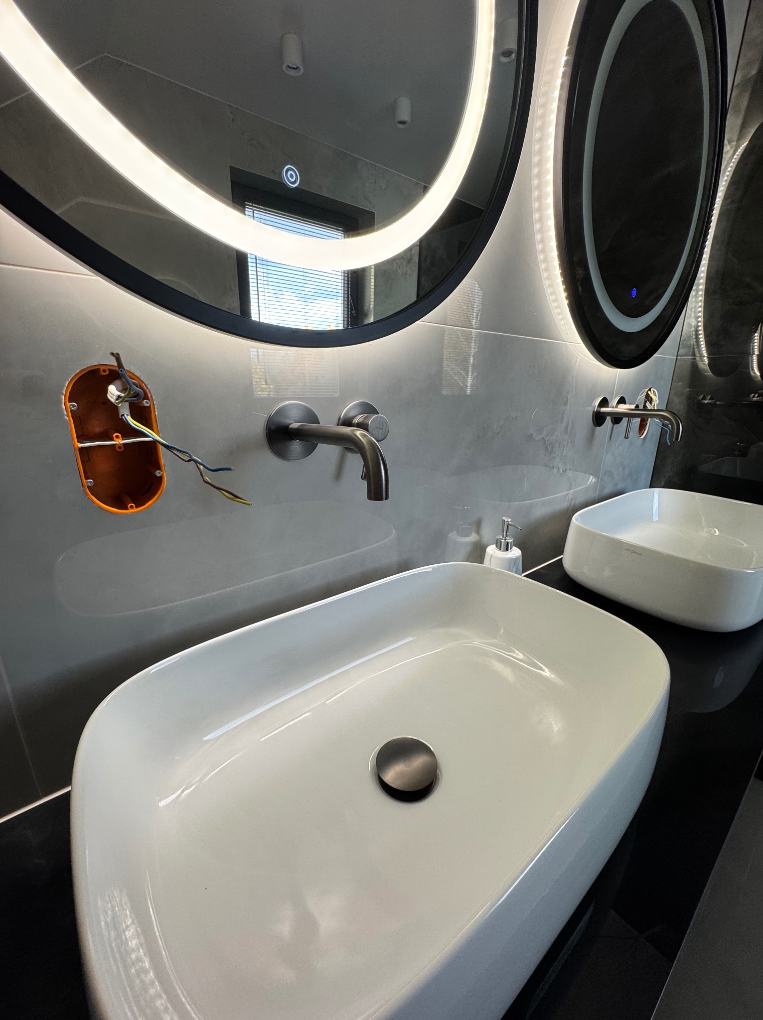 Nowoczesna łazienka z dwiema białymi umywalkami, podświetlanymi lustrami i odsłoniętą instalacją elektryczną. Szare płytki na ścianach.