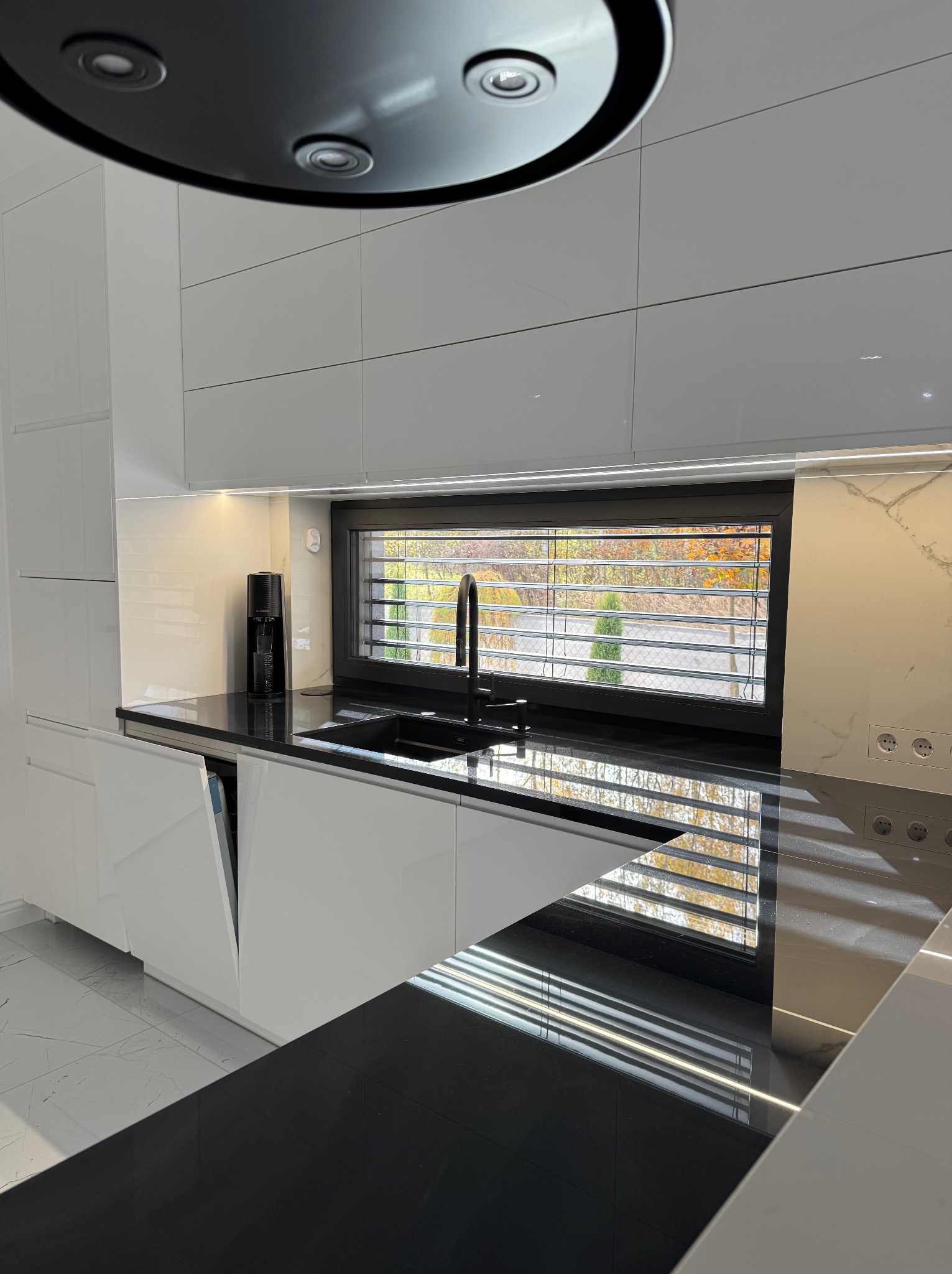 Nowoczesna, biała kuchnia z czarnym blatem i armaturą. Widok na zewnątrz przez okno z żaluzjami. Minimalistyczny design, czystość i porządek.