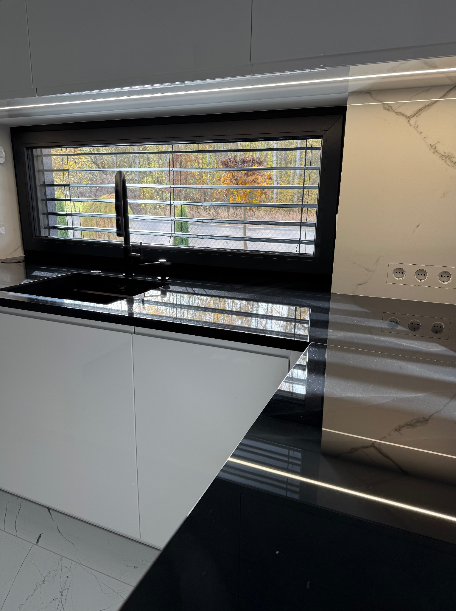 Nowoczesna kuchnia z czarnym blatem i zlewem, okno z żaluzjami w tle. Minimalistyczny design, białe szafki, marmurowa ściana z gniazdkami. Oświetlenie LED podkreśla detale.