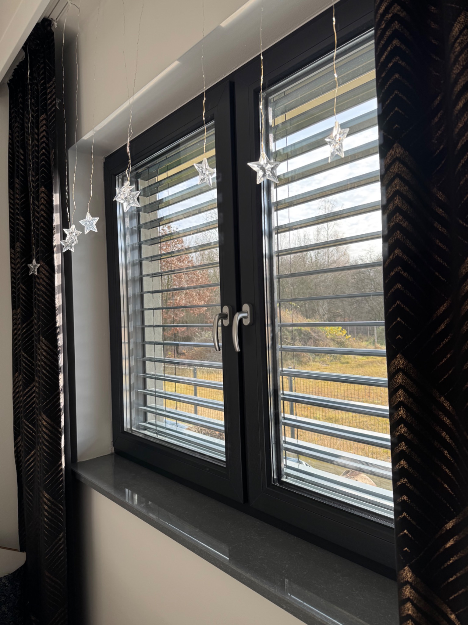 Czarne okno z żaluzjami i dekoracjami w kształcie gwiazd, widok na zewnątrz. Ciemna zasłona z wzorem po lewej stronie. Kamienny parapet.